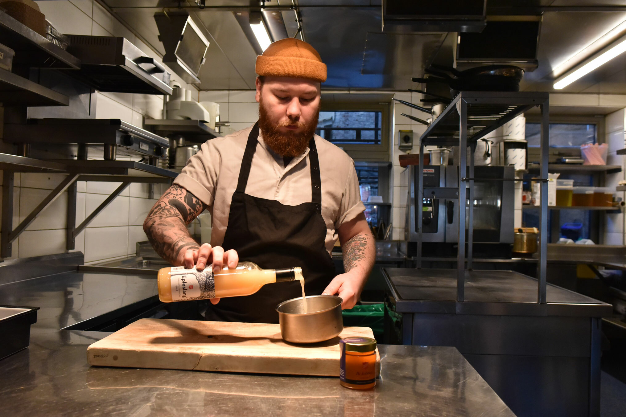 Foto: Mann i lys kokkeuniform med svart forkle over heller eplemost fra en flaske over i en liten kjele. Han har rødt skjegg og oransje lue og på armene har han tatoveringer.