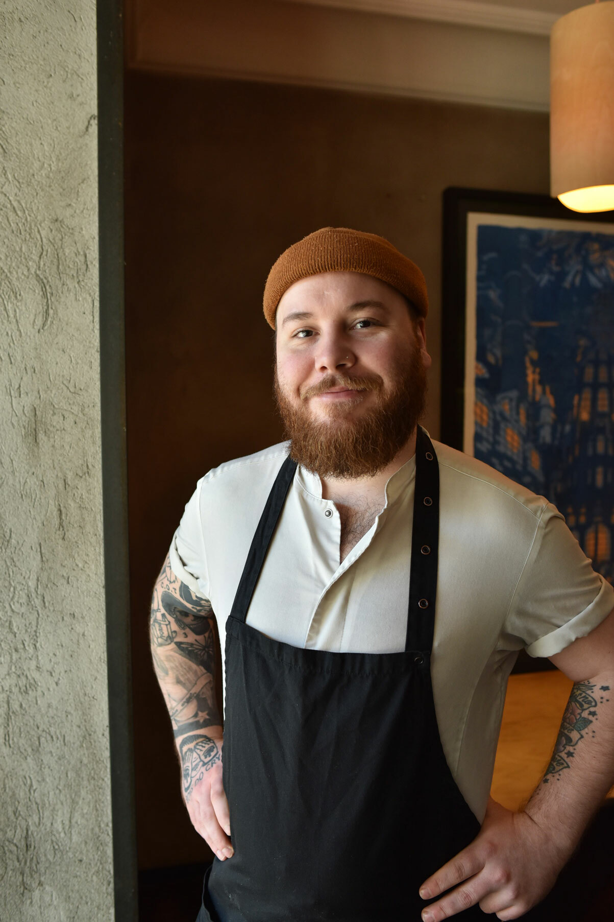 Foto: Mann med rødt skjegg og orange lue har på seg hvit kokketrøye med svart forkle over. På begge armene har han tatoveringer. Han smiler mot kamera.