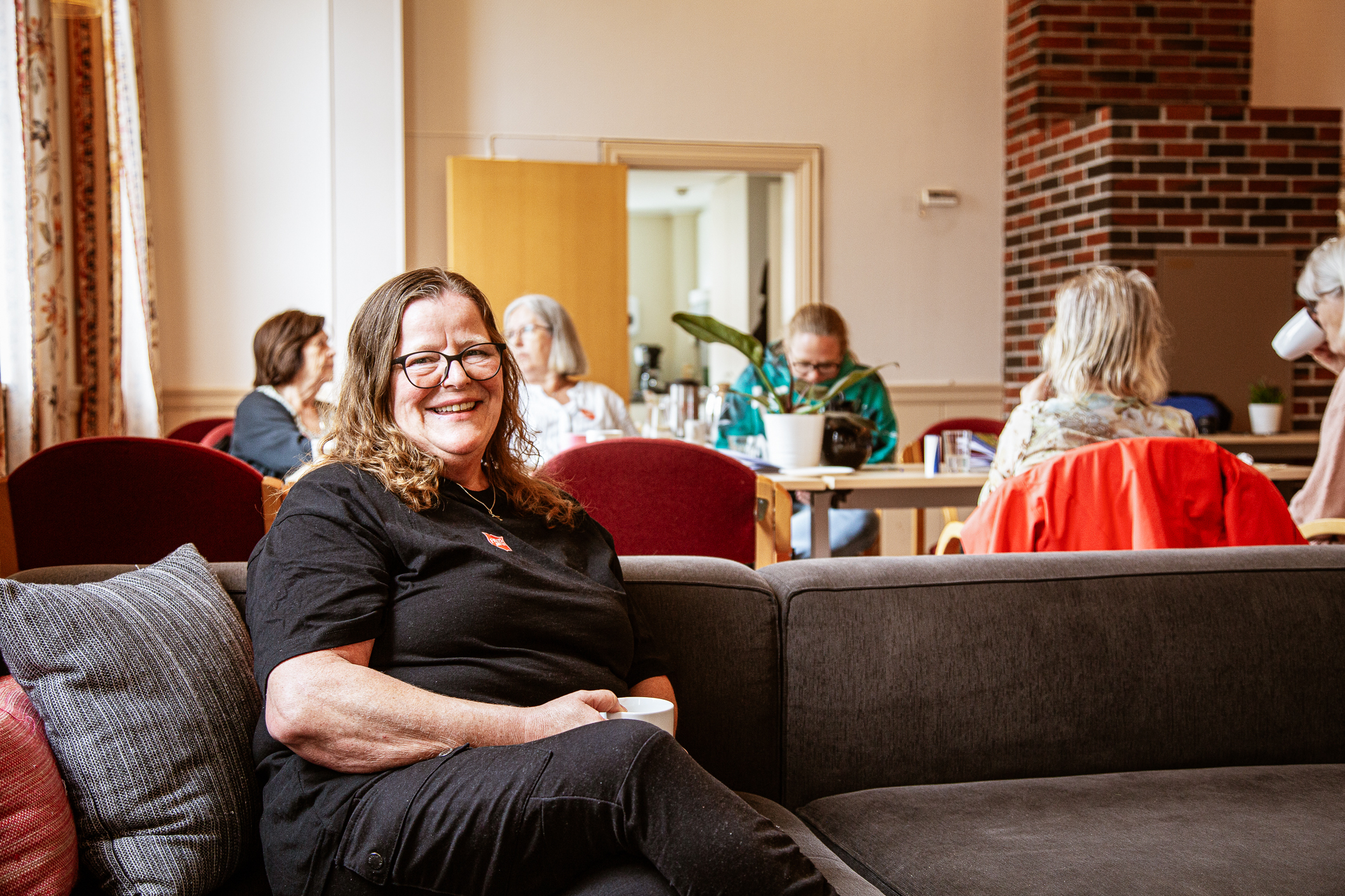 middelaldrende kvinne sitter i en sofa i en menighetssal. Bak henne sitter det flere gjester rundt bordene.