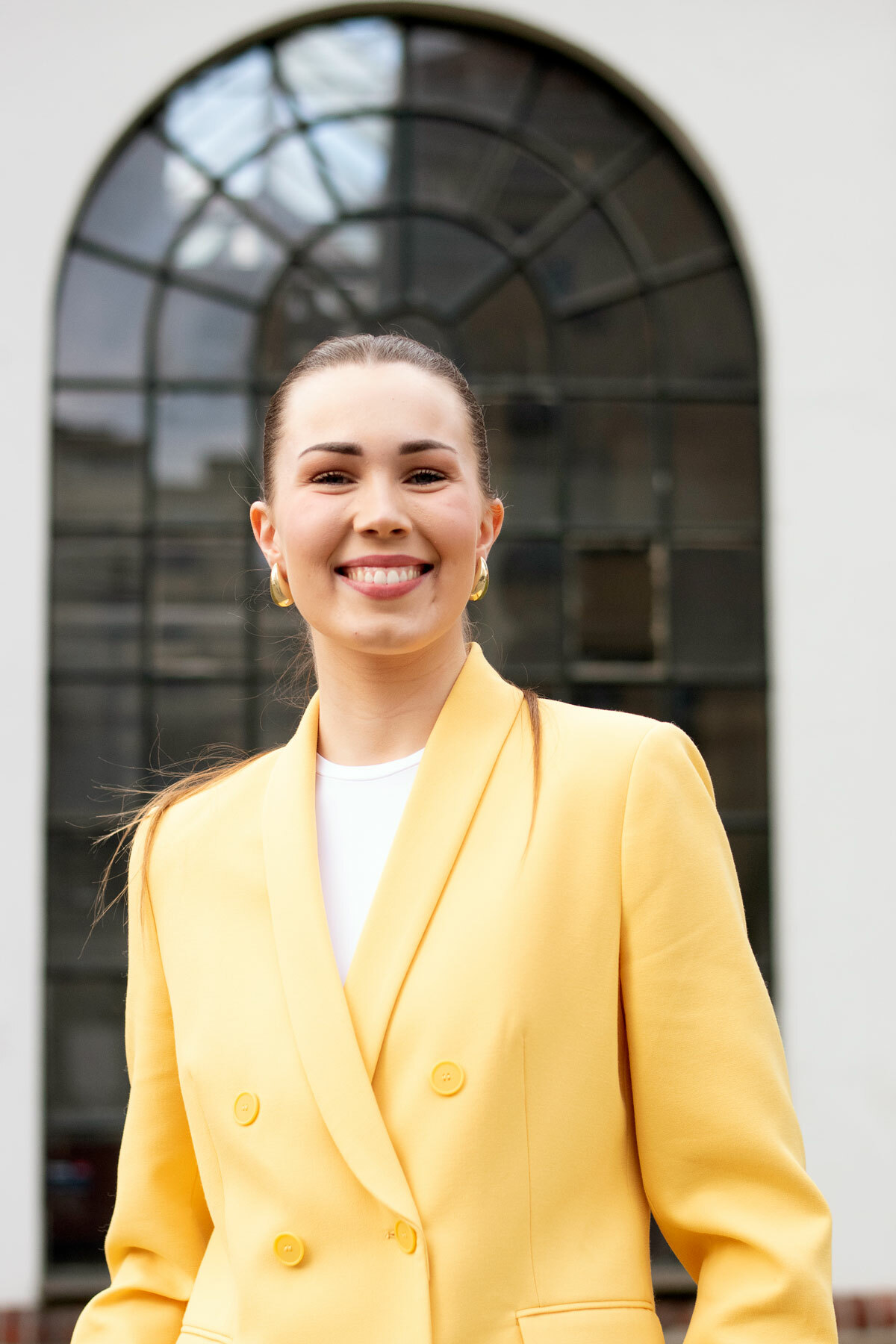 Foto: Ung kvinne med langt, mørkt hår i hestehale smiler foran et rundt vindu. Hun har på seg en gul jakke.