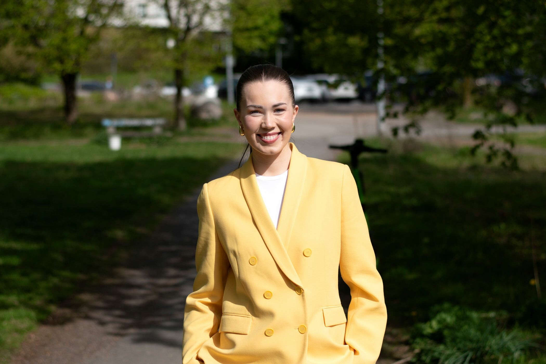 Foto: Ung kvinne med langt, mørkt hår i hestehale smiler i en grønn park. Hun har på seg en gul jakke.