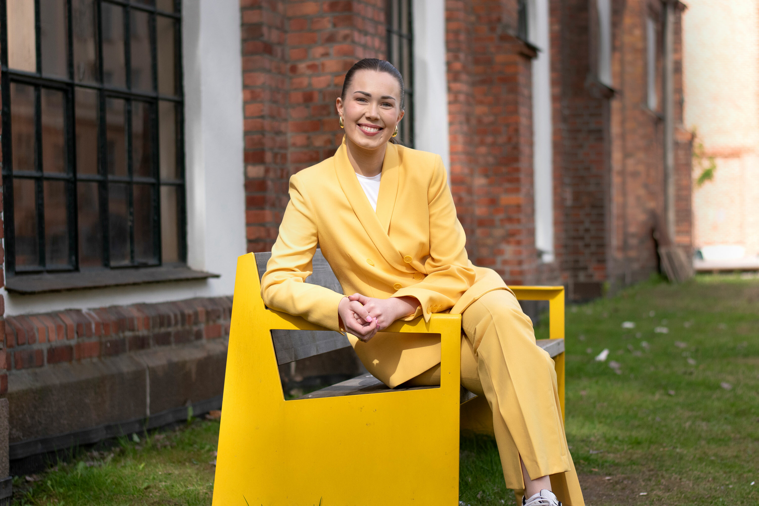 Foto: Ung kvinne med langt, mørkt hår i hestehale sitter på en gul benk i metall. Hun har på seg en gul jakke.