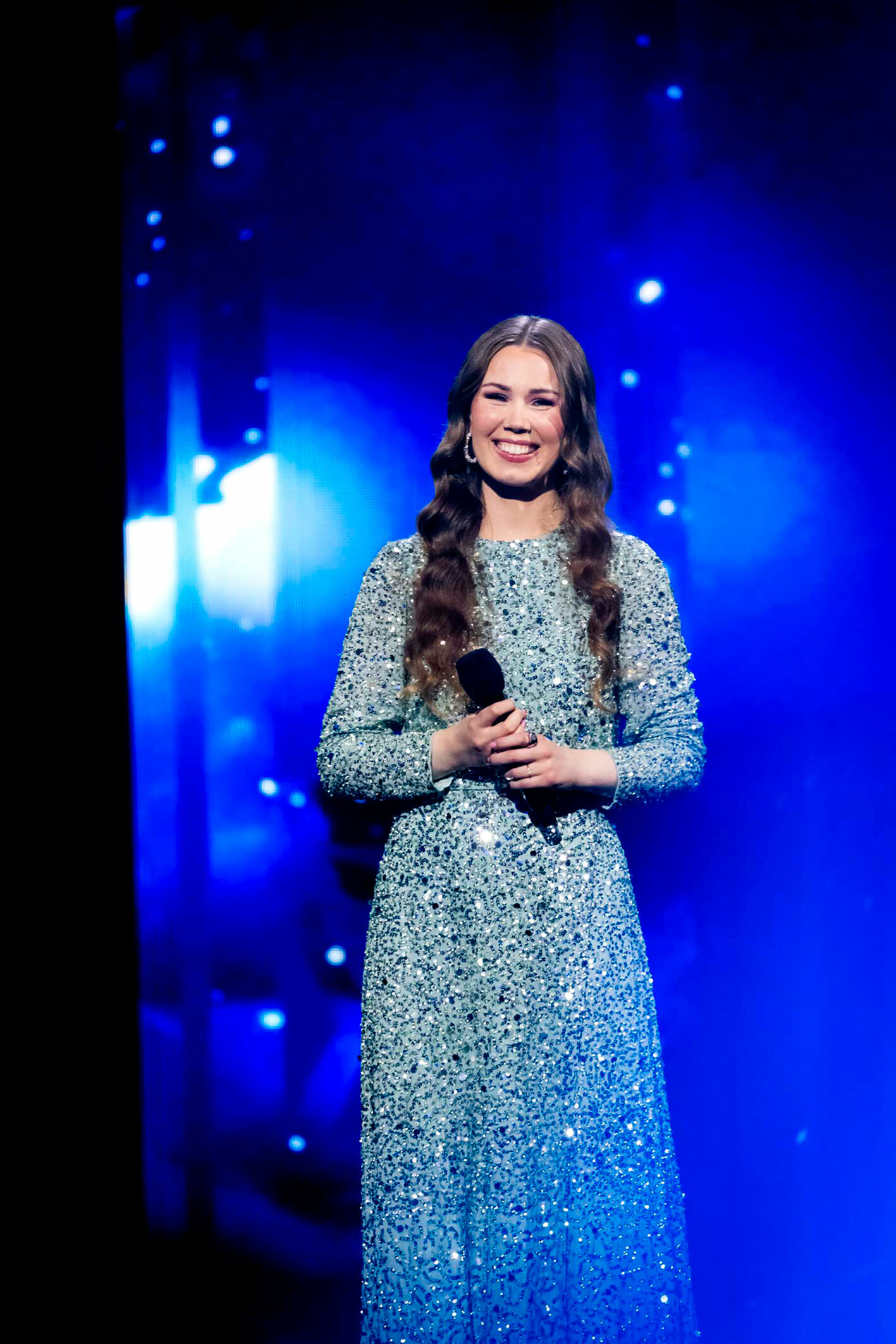 Foto: Foto: Ung kvinne med langt, mørkt hår i hestehale smiler fra en scene. Hun har på seg kjole med paljetter og holder en mikrofon i hendene.