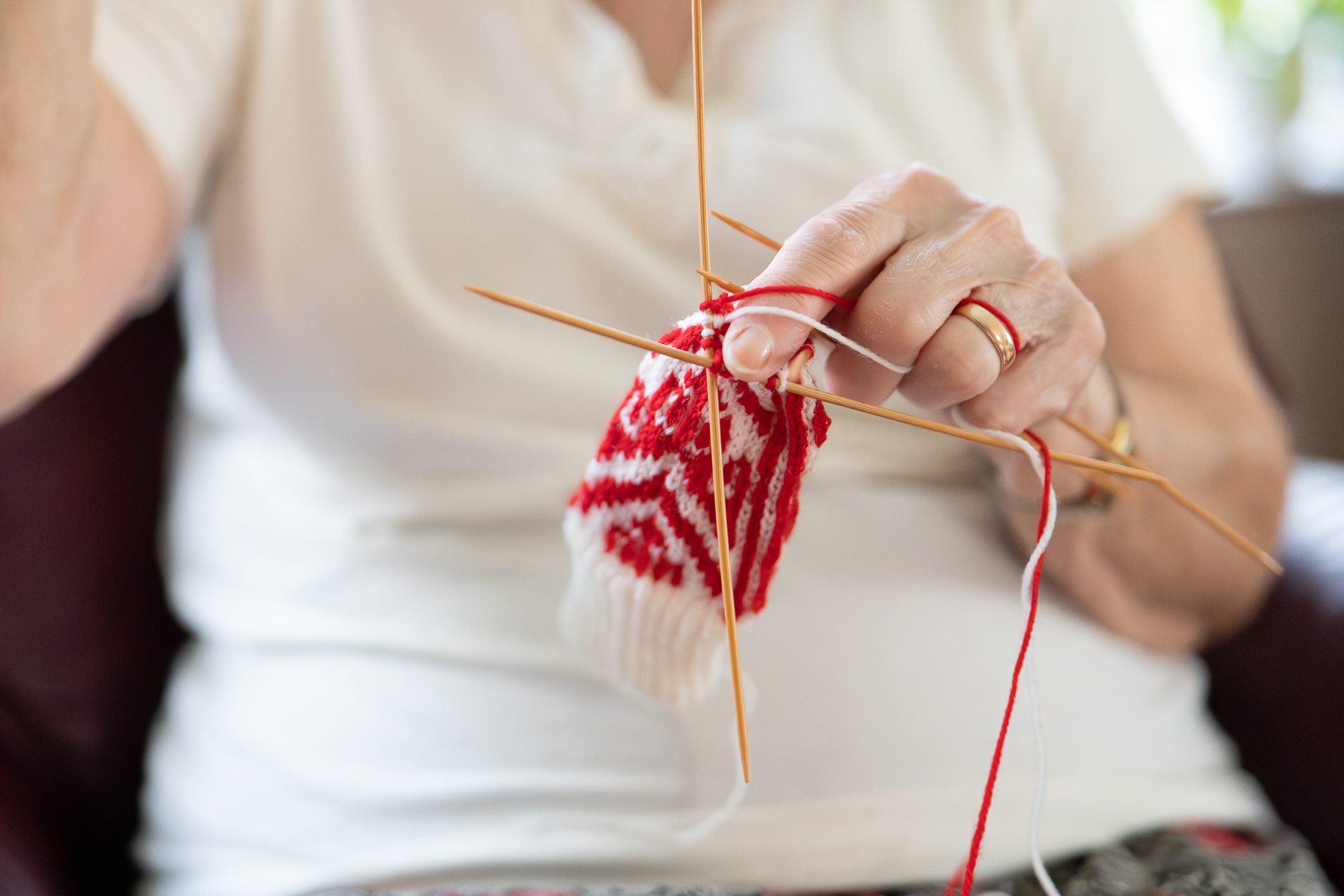 Hender til en eldre kvinne som strikker en bitteliten selbuvott i rødt og hvitt. Hun har en hvit topp på seg.