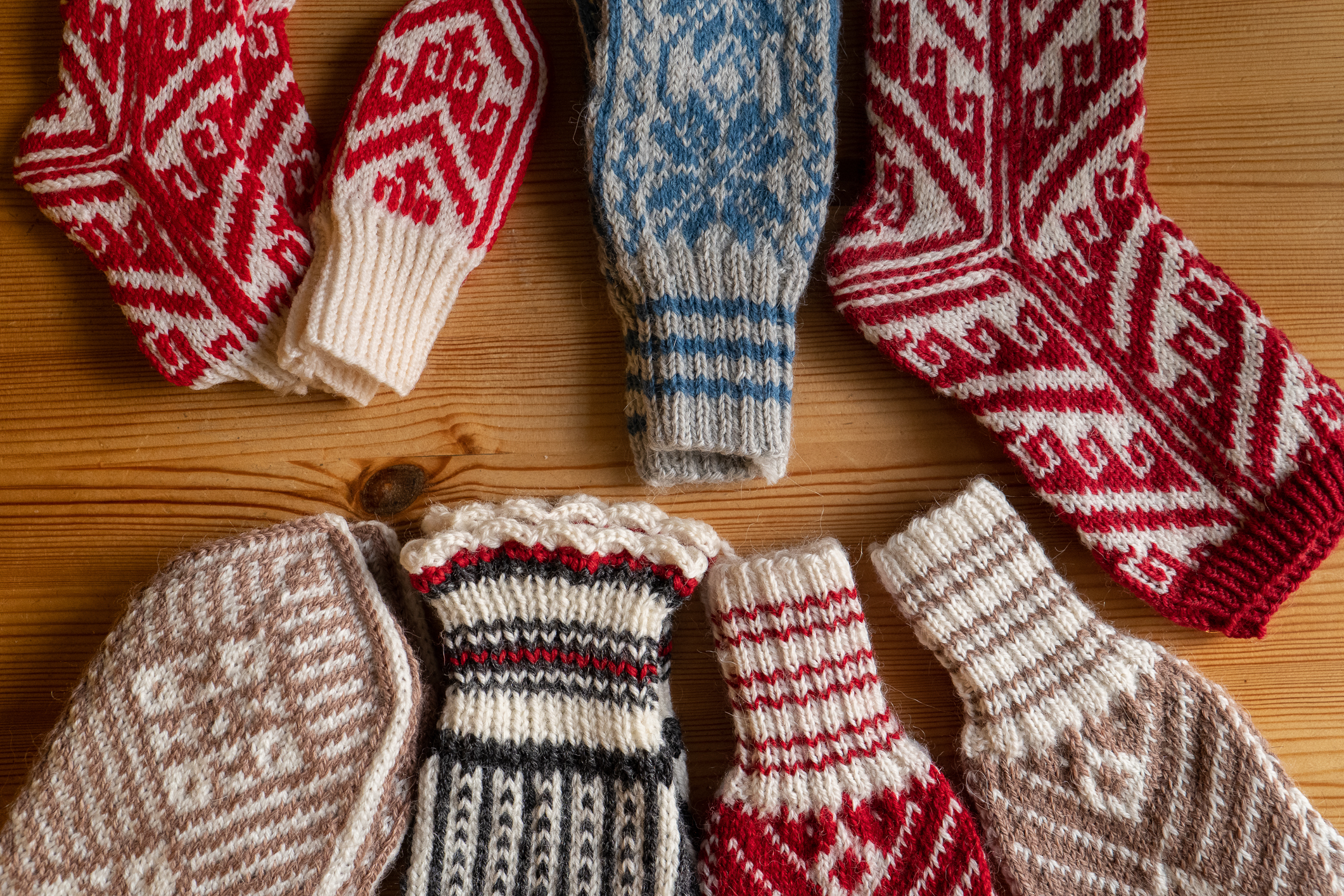 På et trebord ligger åtte par selbuvotter og sokker i vakre farger og mønstre. Mest røde og hvite, men også blå og brå og lysebrune og hvite.