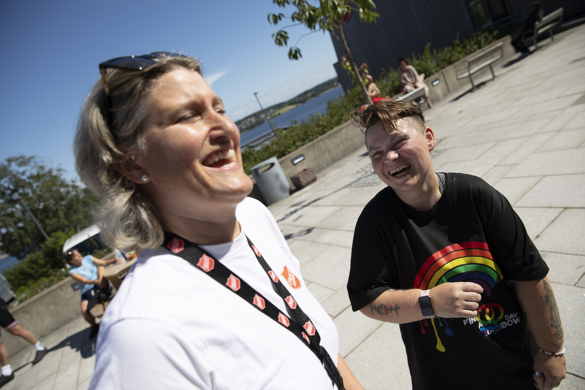 Foto: To kvinner står ute på en åpen plass med havet i bakgrunnen. Begge ler godt.