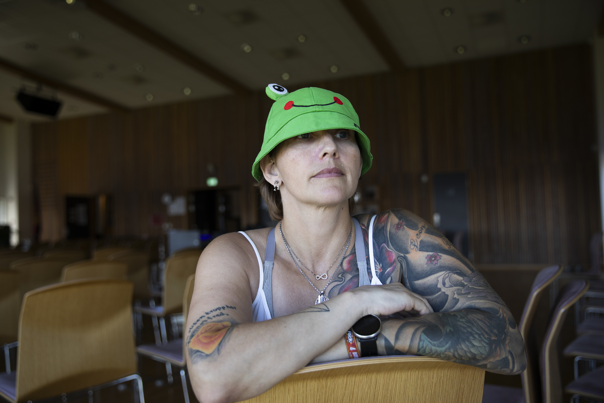 Foto: Kvinne med mange tatoveringer – og grønn froskehatt – sitter feil vei på en stol inne i en sal.