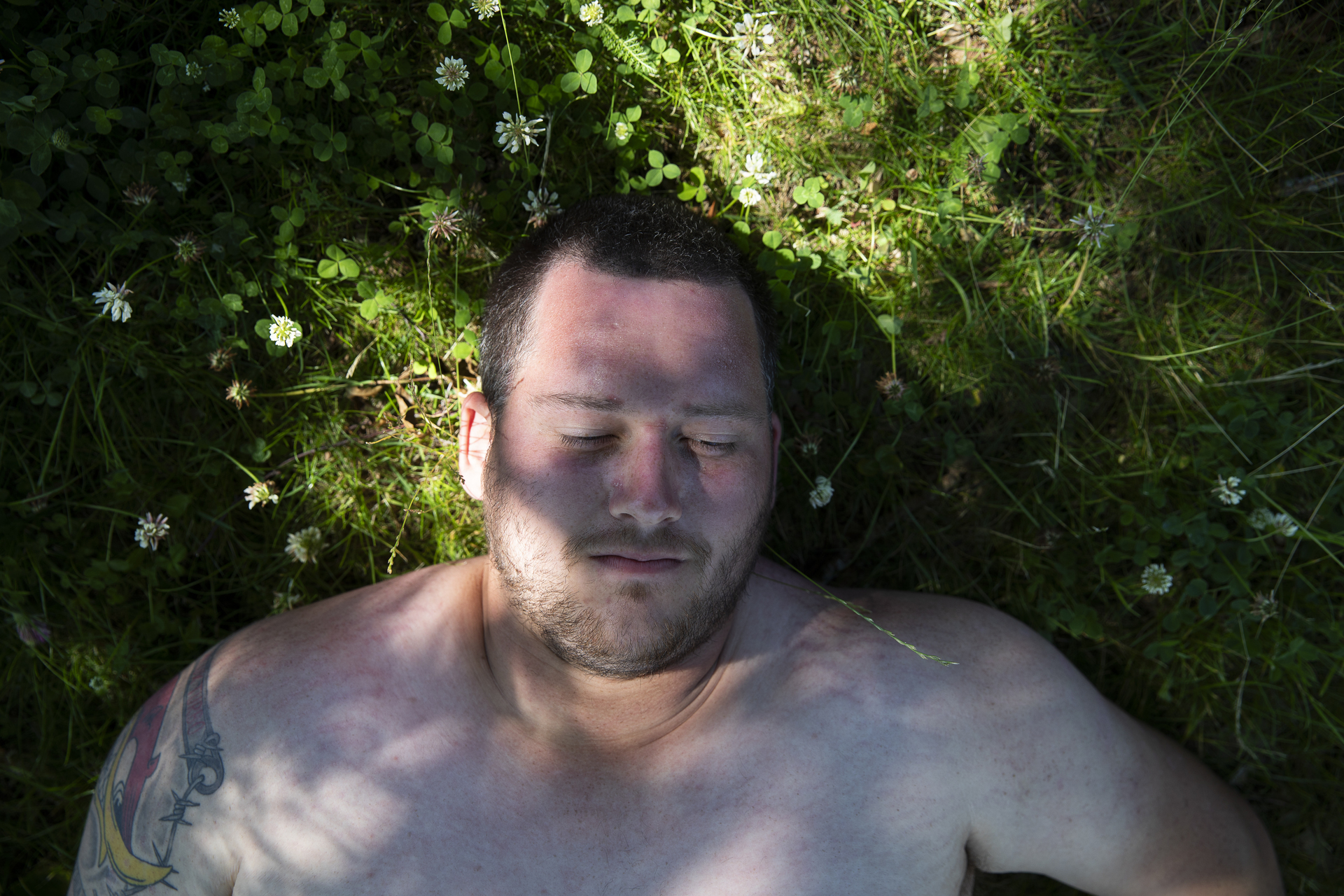 Foto: Man som ligger i gresset med bar overkropp. Vi ser ham bare fra skuldrene og påå. Han har en tatovering på høyre arm-
