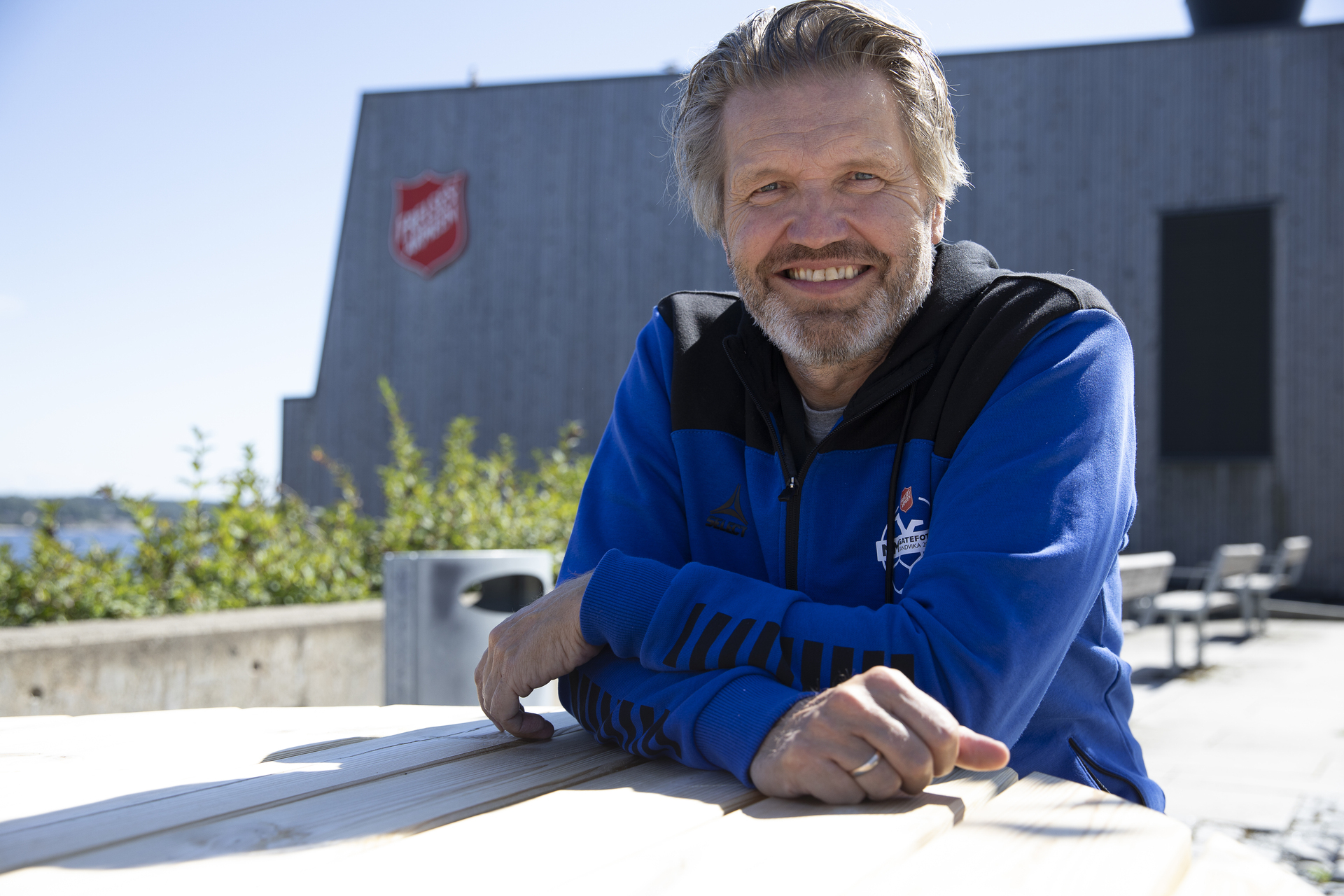 Foto: Mann med skjegg og gråblondt hår sitter ute ved et bord i treningsjakke. I bakgrunnen sees Frelsesarmeens logo på et moderne bygg i tre.