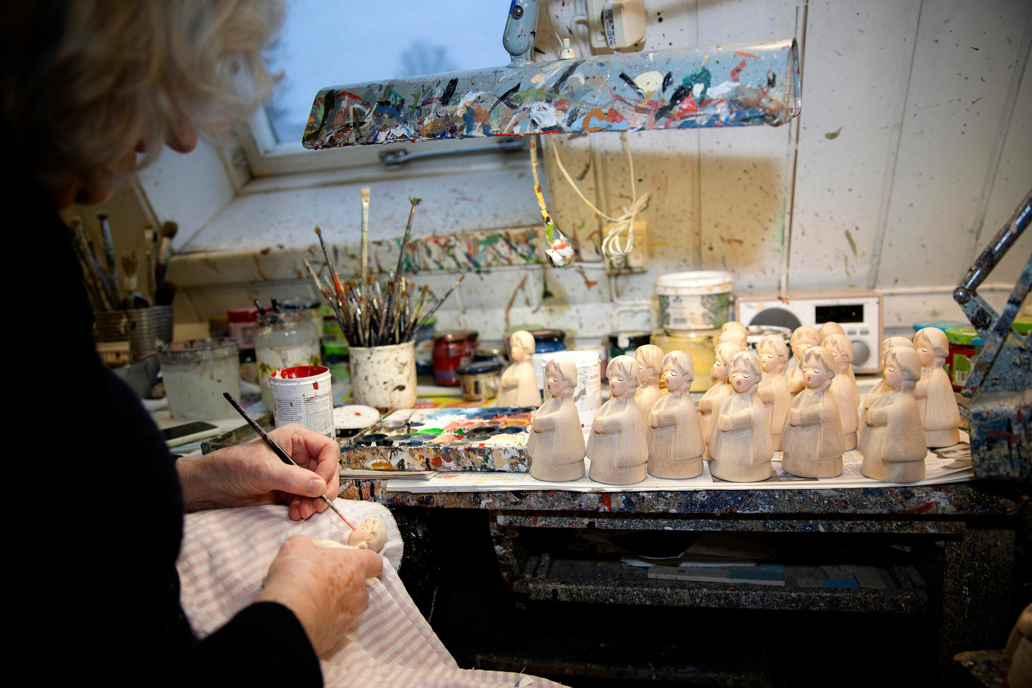 Foto: Kvinne i svart genser med grått hår maler på et helt brett med engler. På bordet står en rekke pensler.