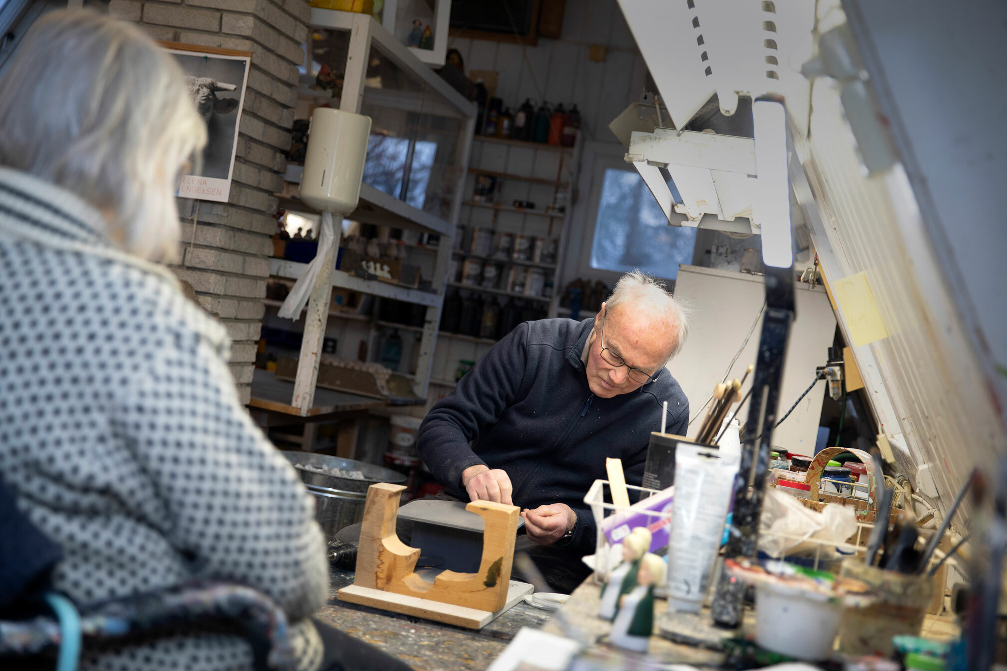Foto: Kvinne med grått hår i hvit og svart ullgenser sitter i en stol ved et arbeidsbord. På den andre siden av bordet holder en mann med briller konsentrert på med noe.