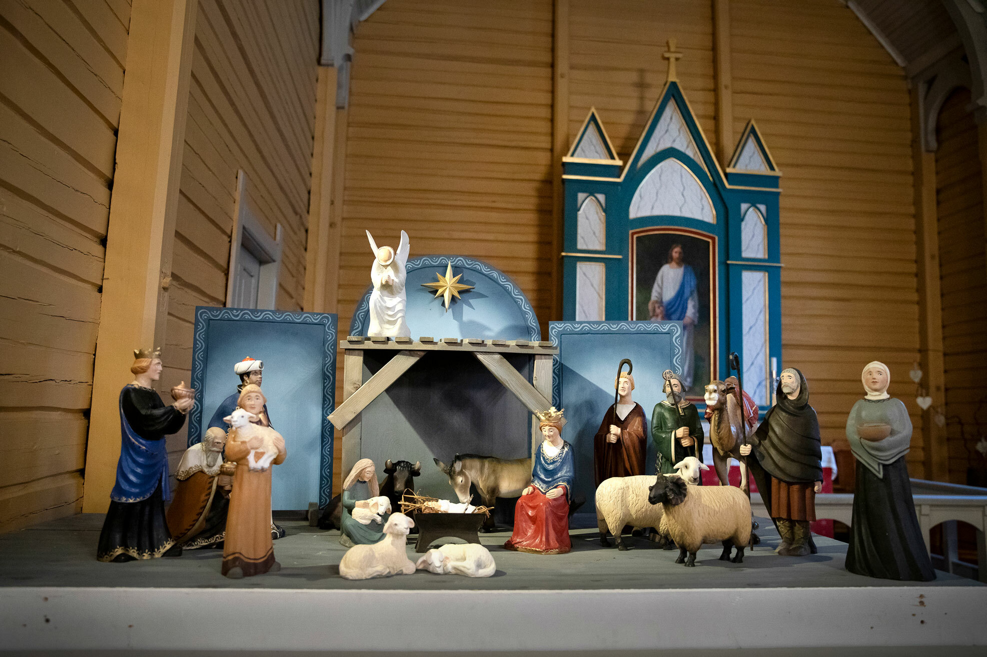 Foto: En stor julekrybbe i tre står utstilt i en kirke. Bak ser vi altertavlen med et bilde av Jesus. Veggene i trekirken er malt gule.