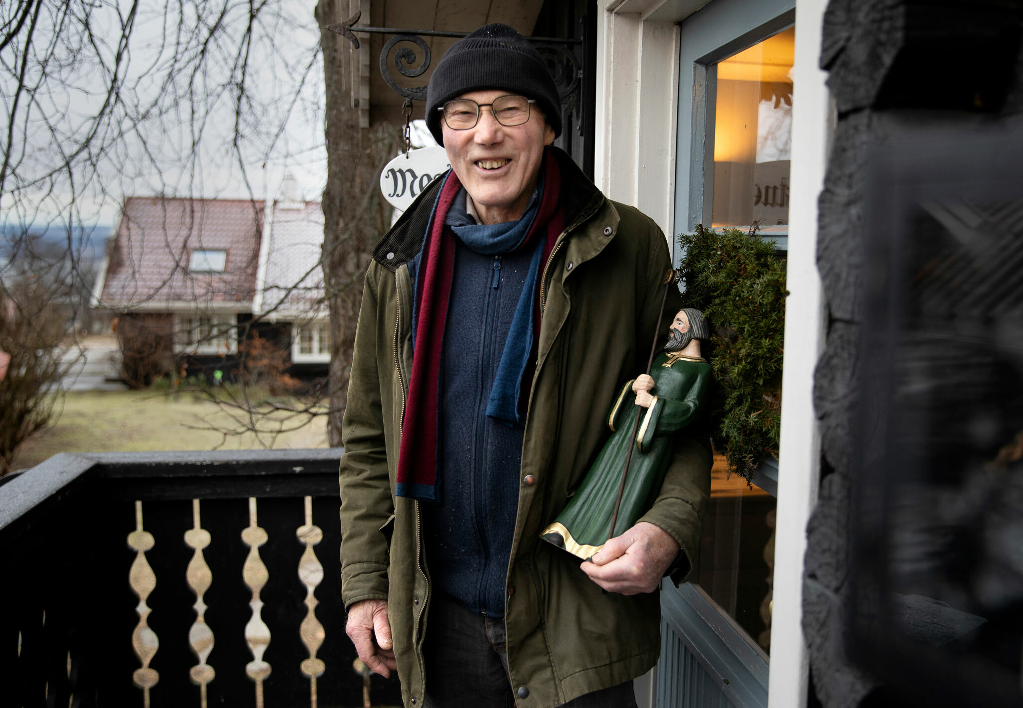 Foto: Eldre mann i mosegrønn jakke står ved inngangsdøra til et verksted. Han har briller og lue på. I venstre armkrok står en statue av en mann i grønn kappe med en stav.