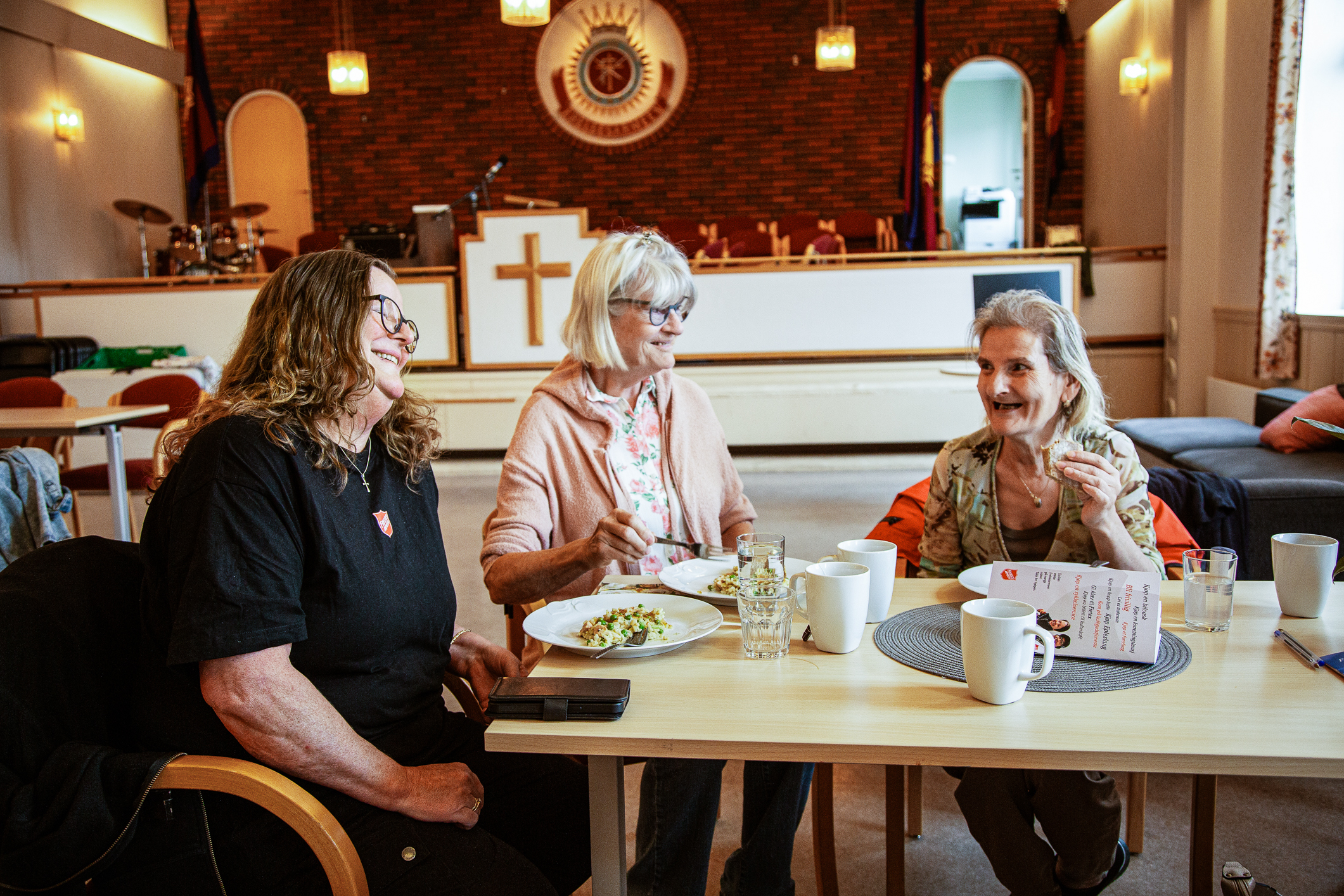 Tre kvinner sitter rundt et bord og spiser i et menighetslokale. i bakgrunnen ser vi en talerstol og Frelsesarmeens skjold.