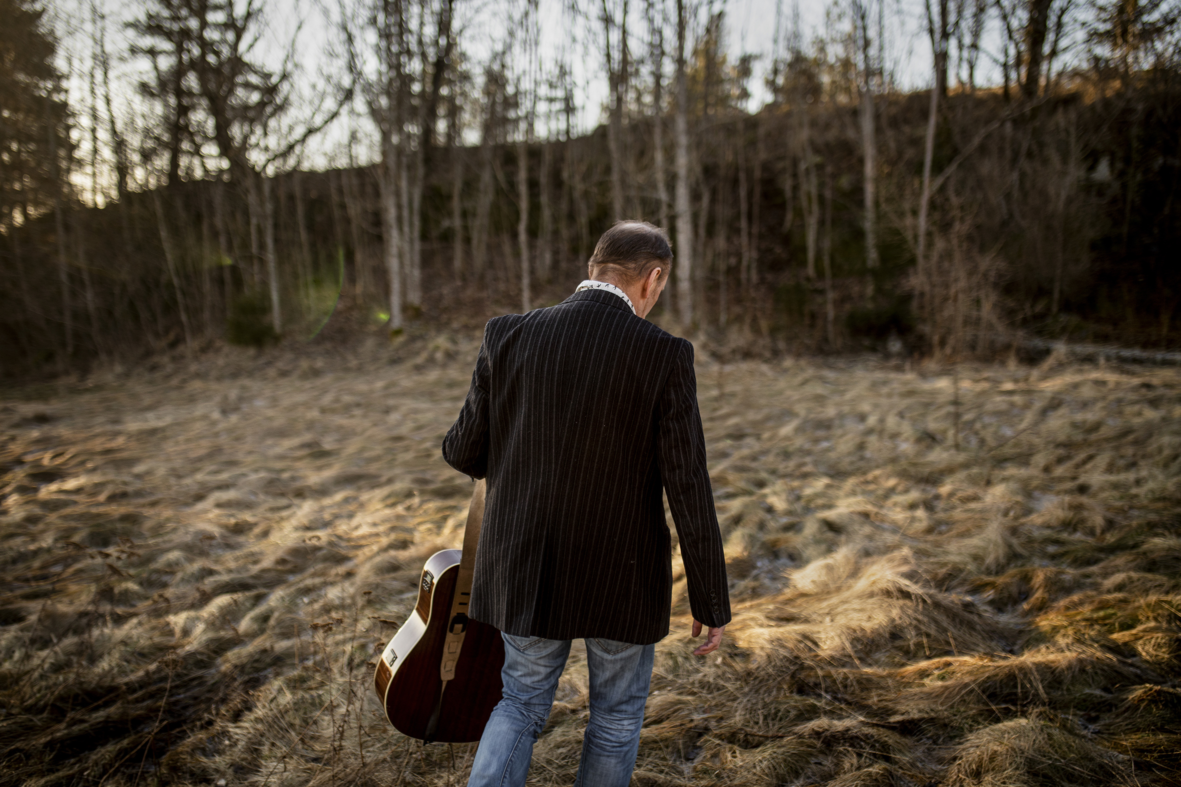 Bildet viser Steinar Hulback på en åpen plass i skogen. Han holder gitaren sin og er på vei bort fra kamera.