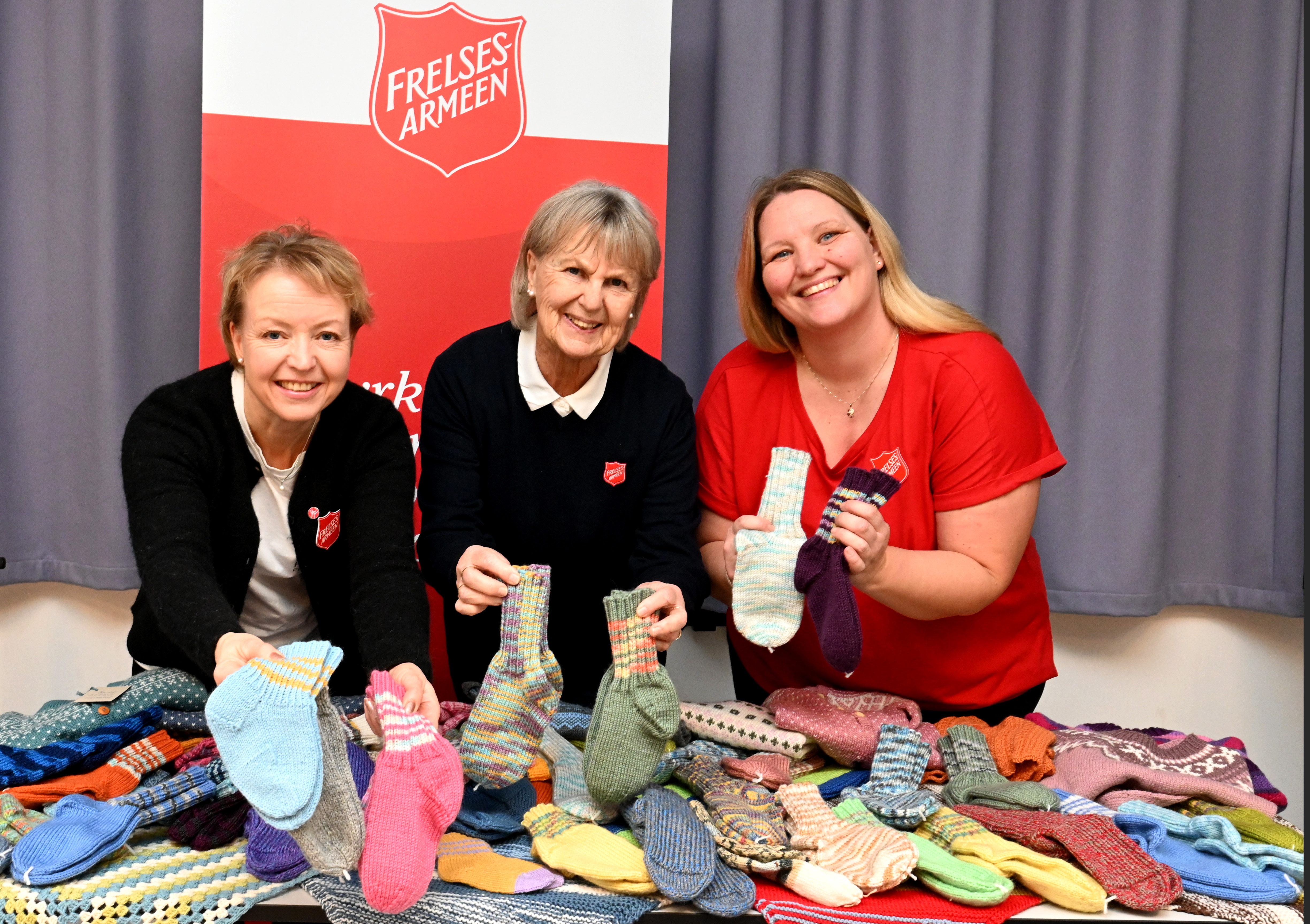 Tre kvinner med frelsesarmeens klær foran et fullt av nystrikkede ullsokker