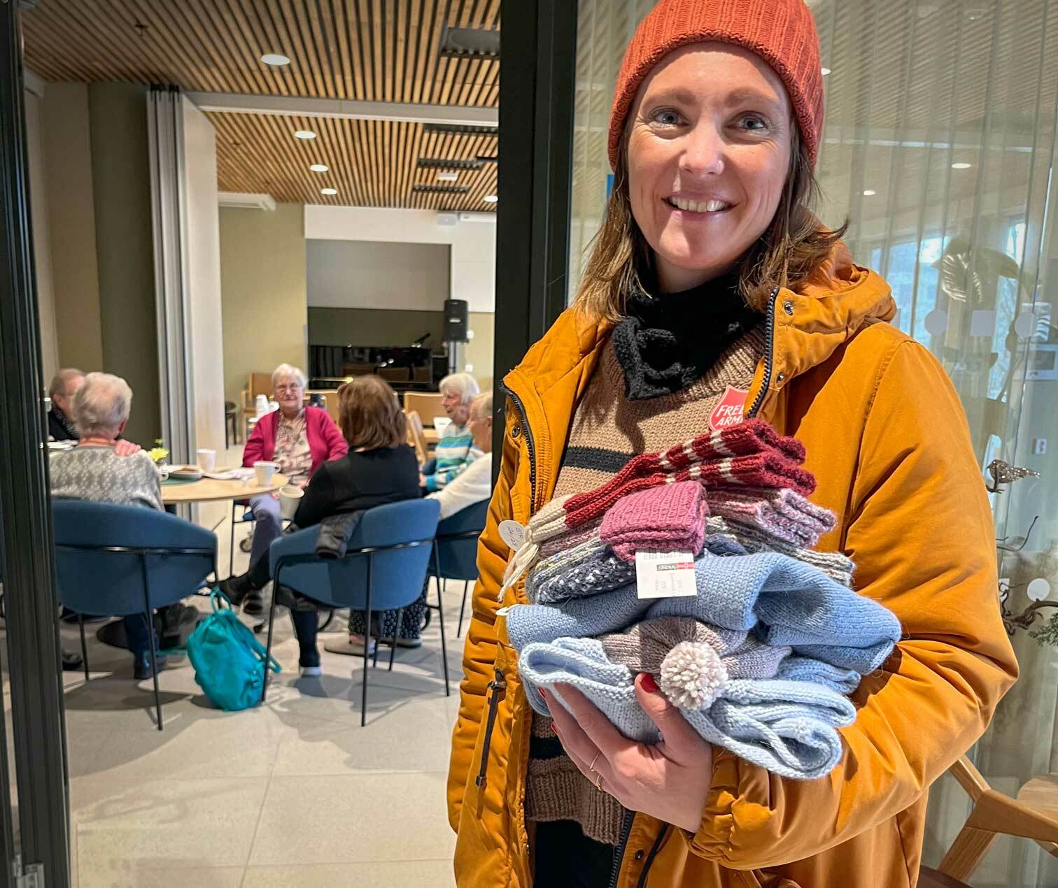Kvinne står med en bunke strikkeklær til barn. I bakgrunnen sitter noen eldre kvinner rundt et bord på et eldresenter.
