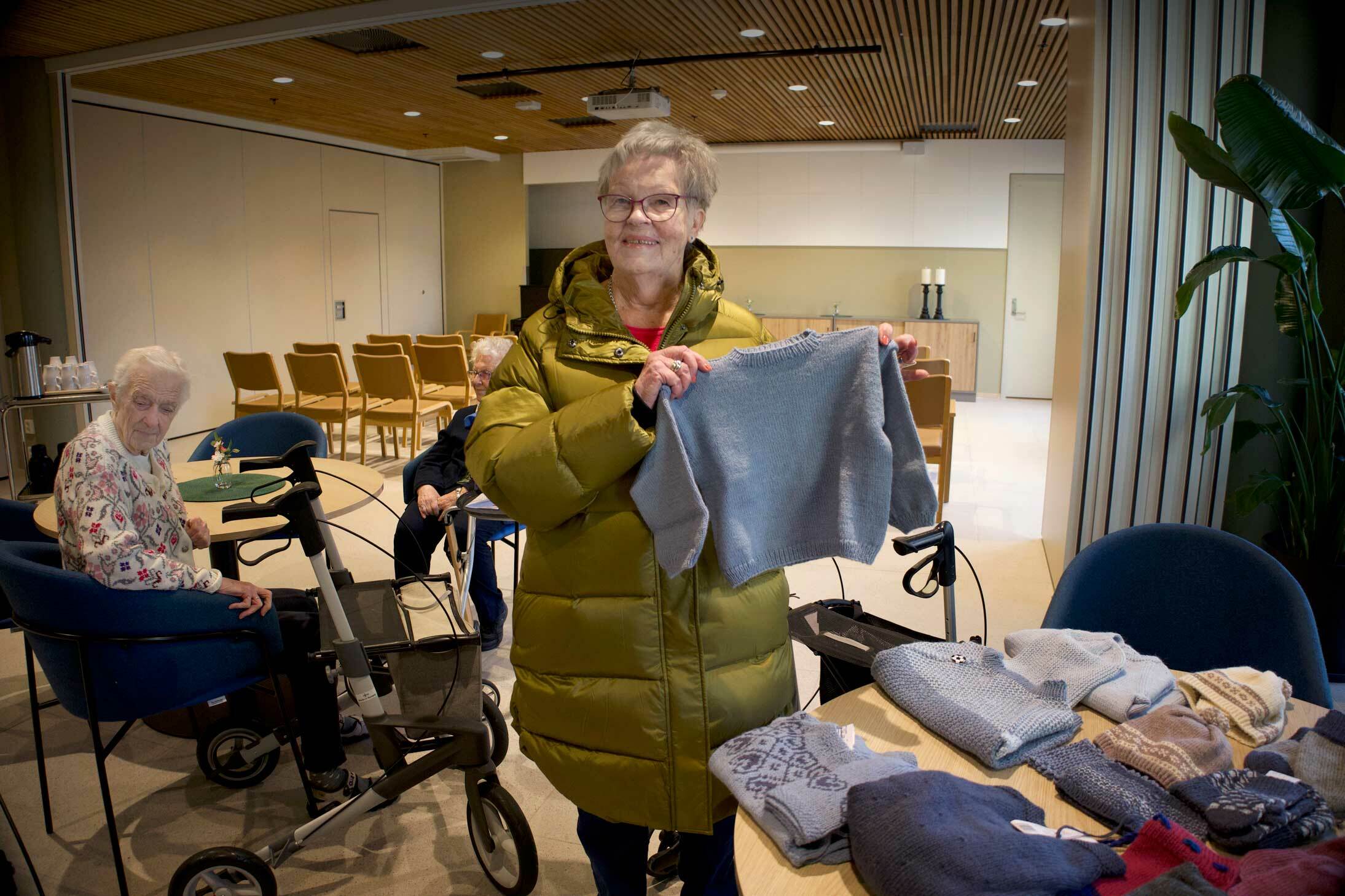 En eldre kvinne med boblejakke holder opp en strikkegenser. På bordet foran henne ligger mange strikkeplagg til barn. En eldre kvinne sitter på et bord ved siden av.