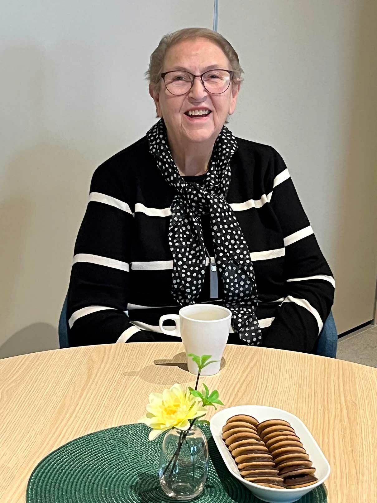 Smilende eldre kvinne med briller sitter ved et bord med kaffekopp og småkaker.