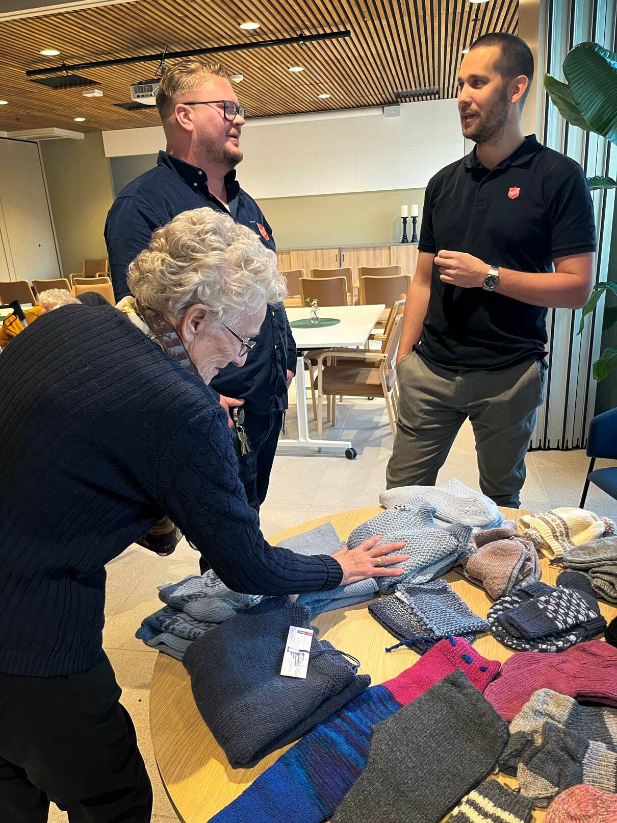 Eldre kvinne kjenner på noen strikkeplagg som ligger på et bord. To menn står