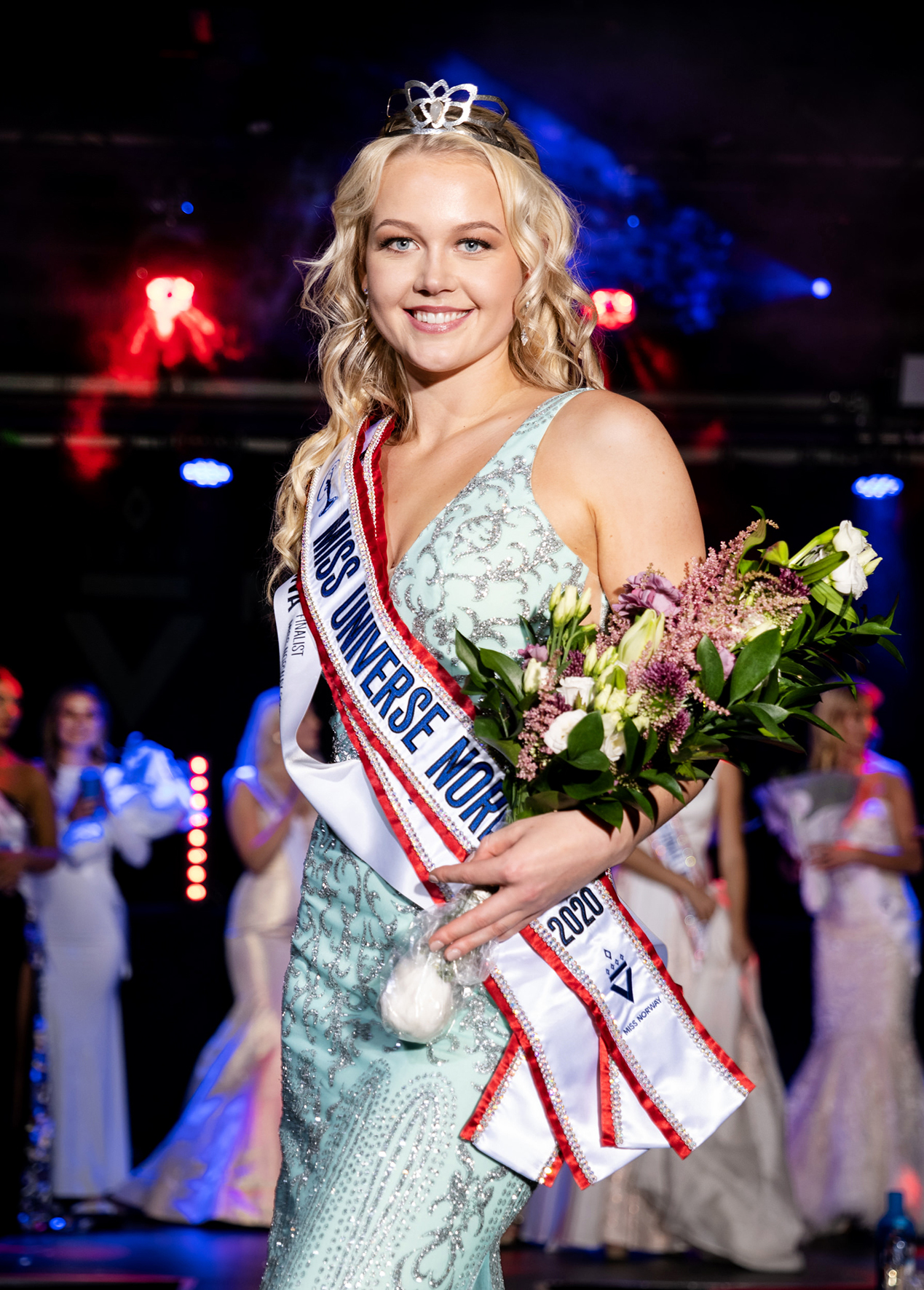 Bildet viser Sunniva på scenen etter at hun har vunnet, hun holder en blomseterbukett og smiler, og har på seg en lyseblå kjole med sølvglitter