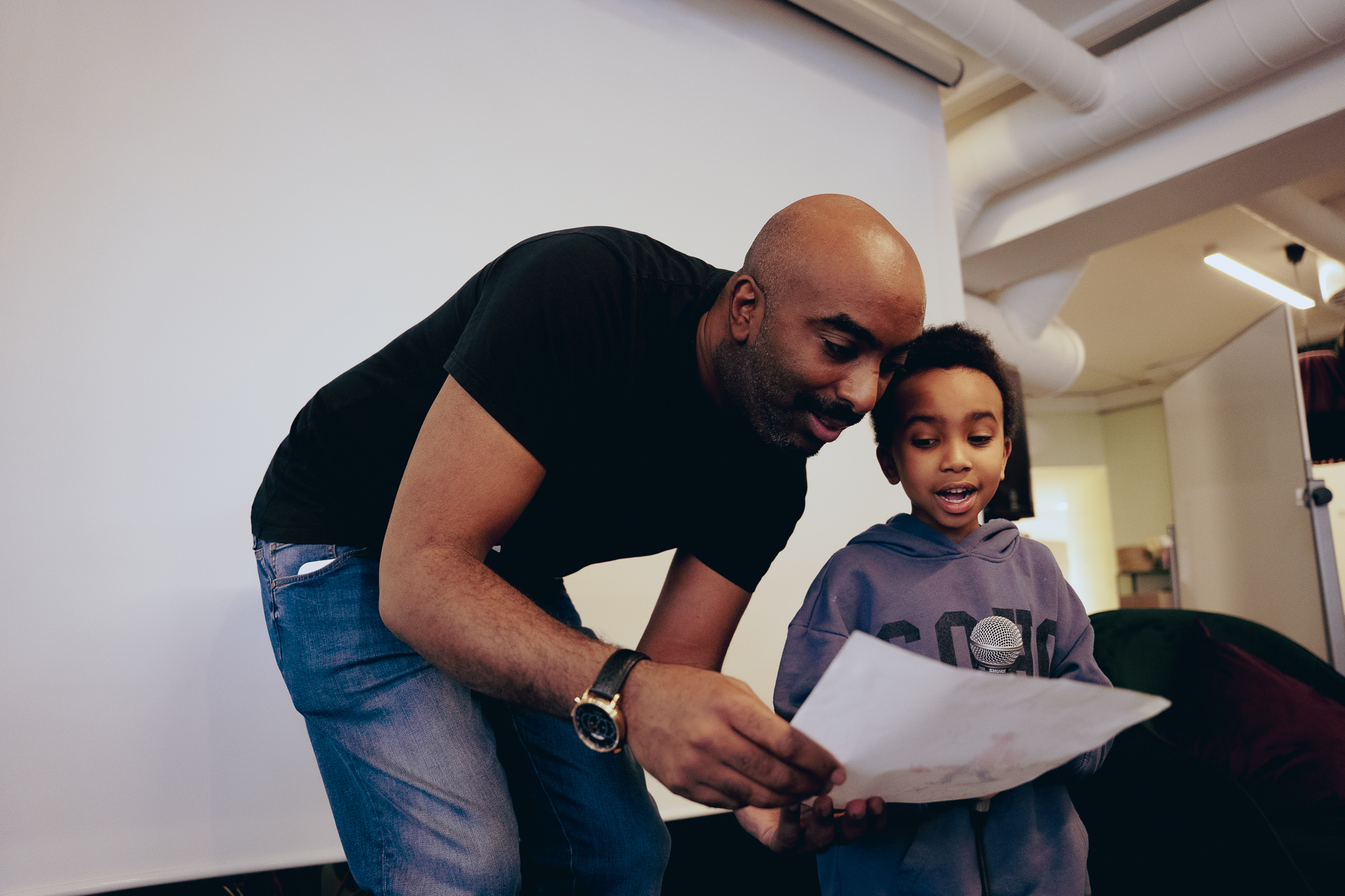 En Mørkhudet og skallet mann med sort t-skjorte smiler mens han hjelper en liten gutt med å lese opp fra et ark som gutten har tegnet på.