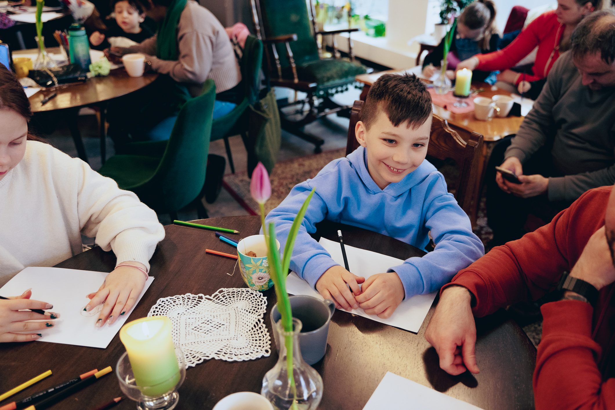 Mange barn sitter rundt flere bord i et koselig lokale. Barna har tegnesaker og ark på bordet. En smilende gutt med lyseblå collegegenser sitter ved ett av bordene og tegner. Han smiler