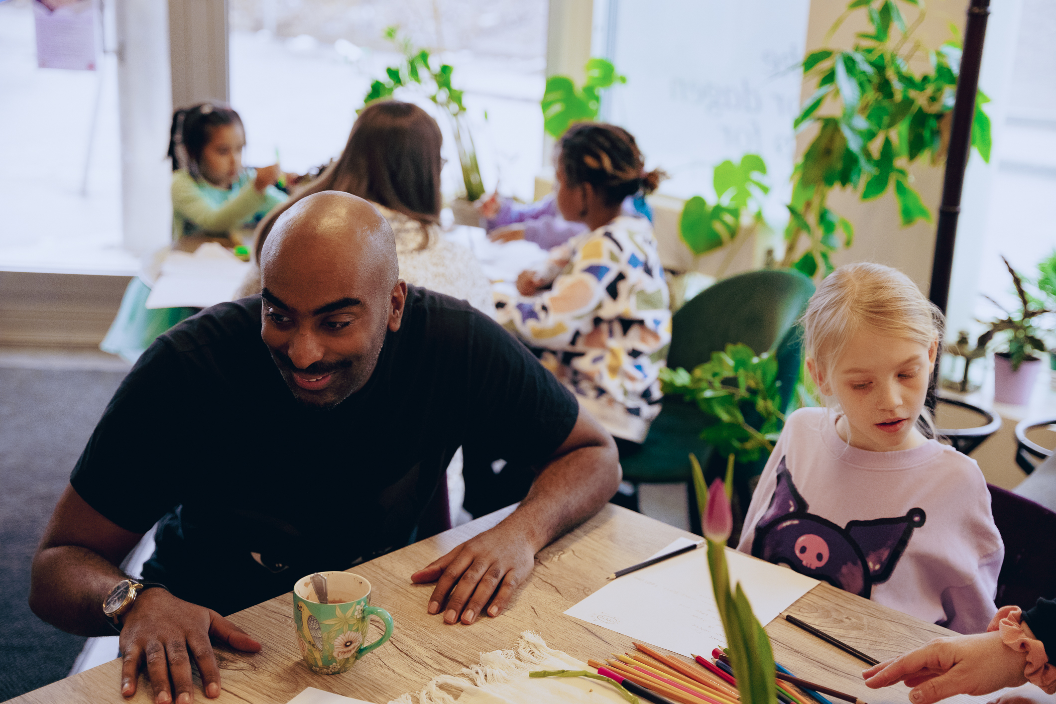 Mange barn sitter rundt flere bord i et koselig lokale. Barna har tegnesaker og ark på bordet. En Mørkhudet og skallet mann med sort collegegenser sitter ved ett av bordene og smiler