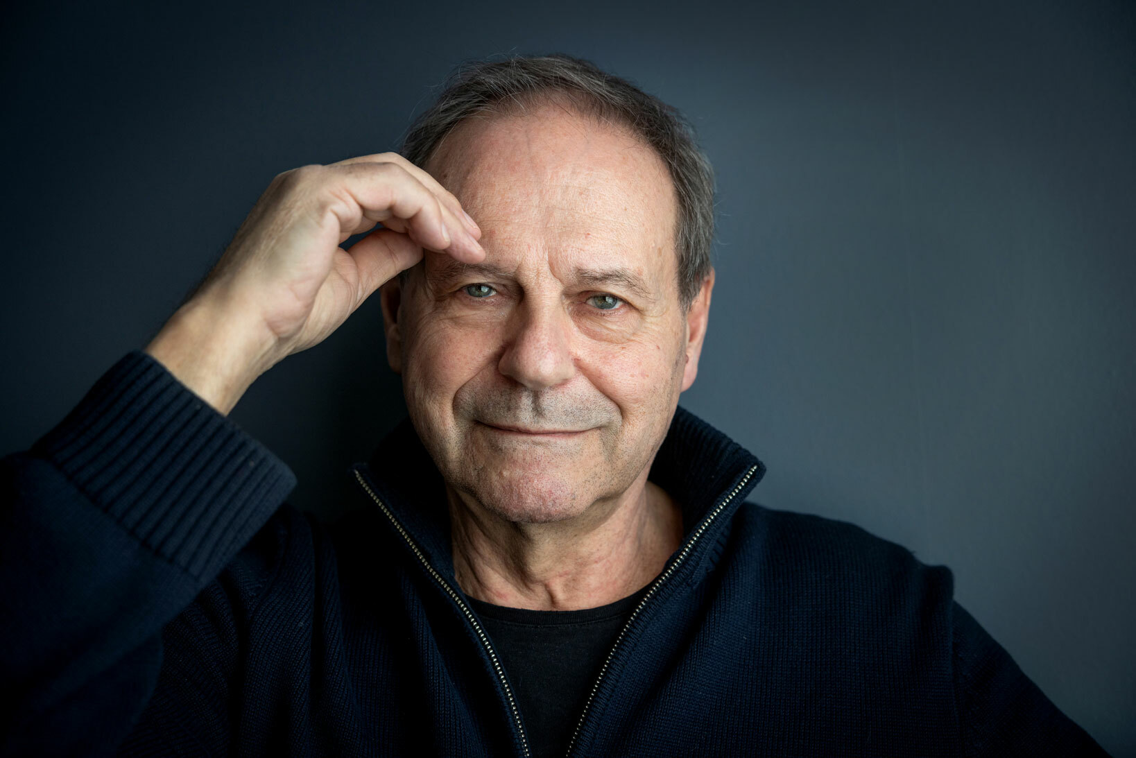 Foto: Mann i 60-årene med mørkeblå genser holder en hånd opp mot pannen. Ser i kamera.