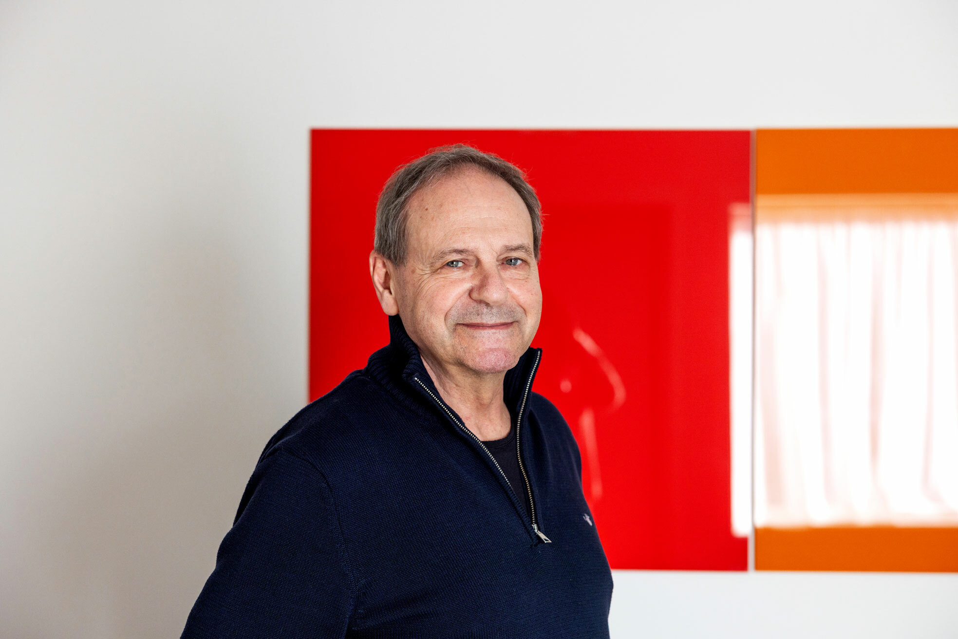 Foto: Mannen i 60-årene med mørkeblå genser ser i kamera. Han står foran en rød, rektangulær glassplate på veggen.