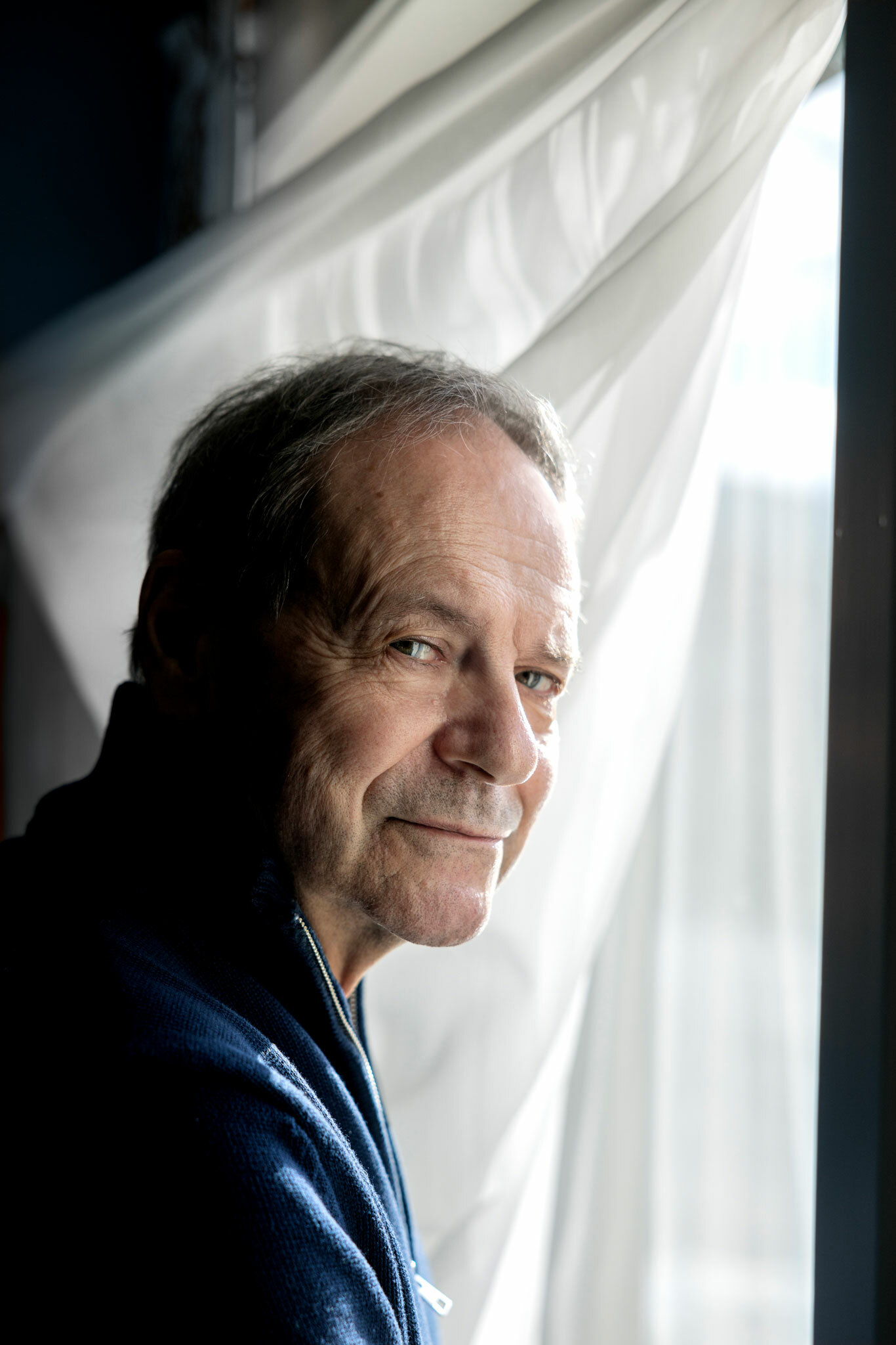 Foto: Mannen i 60-årene med mørkeblå genser ser i kamera. Han står foran en hvit gardin i et vindu.