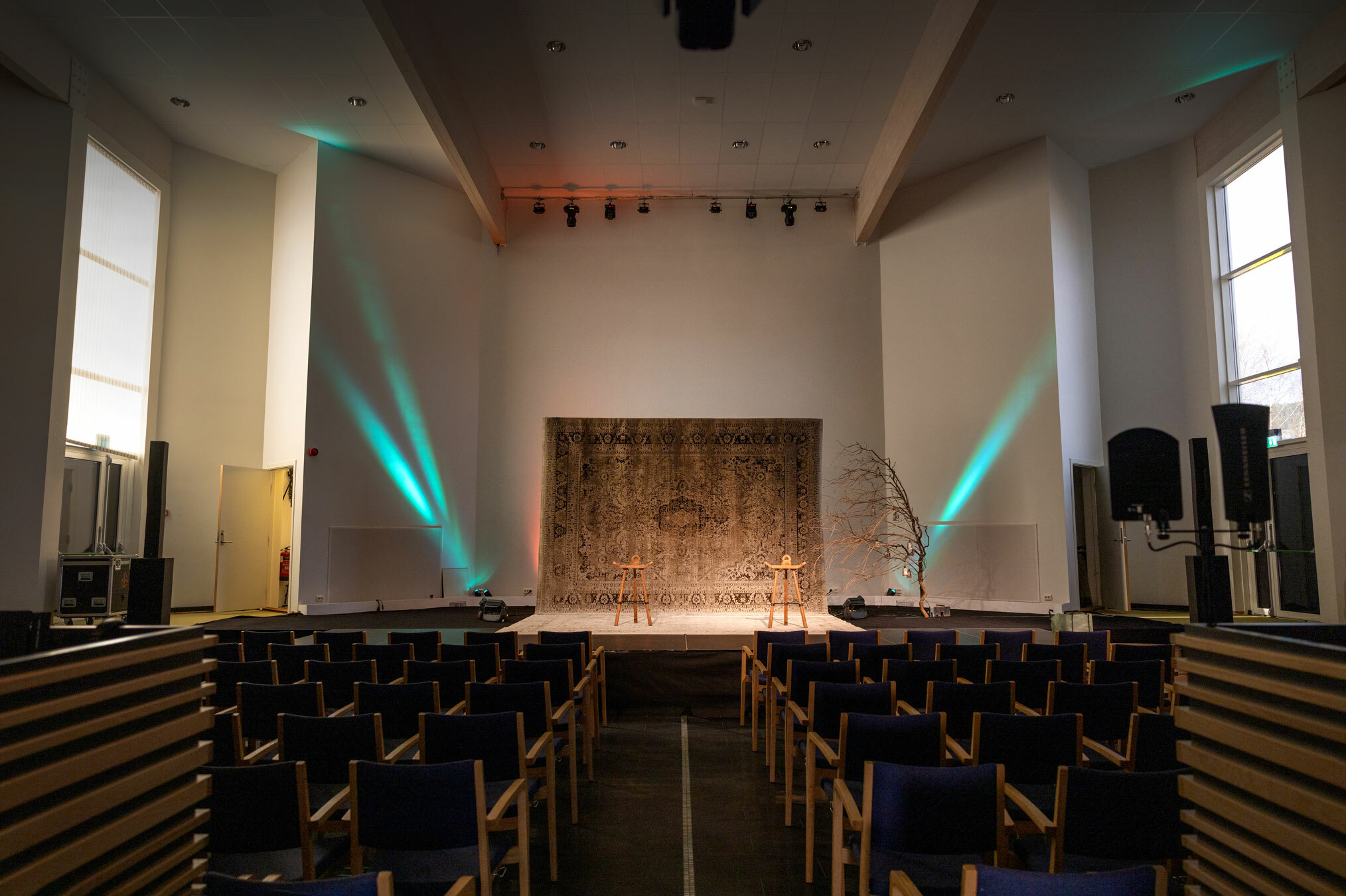 Foto: Av en stor sal med mange stoler og en scene i enden. Der henger det et teppe i enden. Her er det grønne lyskastere oppover veggen.