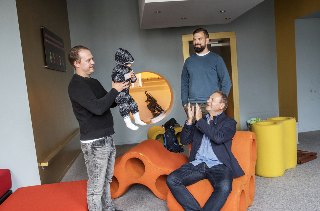 Foto: Far i 60-årene med to sønner i 30-årene og et barnebarn som er baby. De er i et fargerikt rom med mange puffer og andre former.