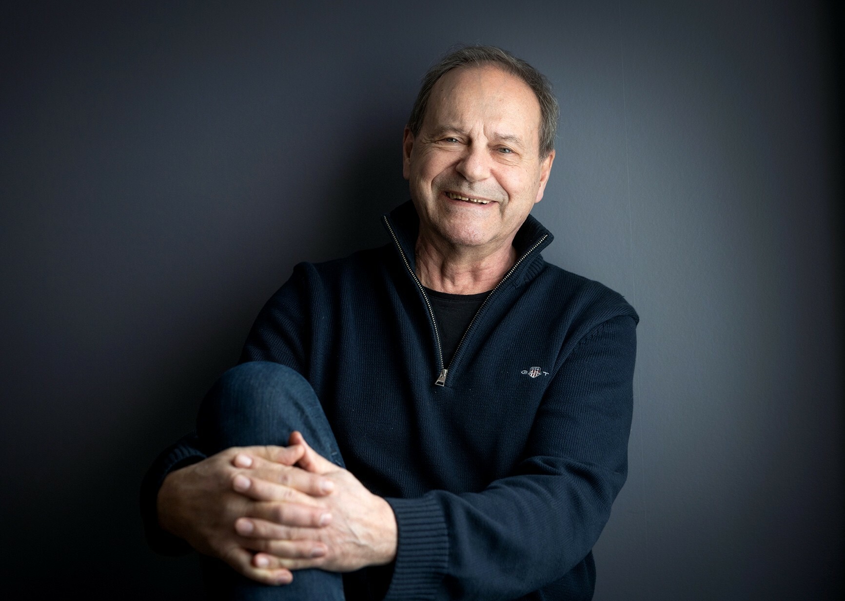 Foto: Mann i mørkeblå genser sitter avslappet inn mot en mørkeblå vegg. Han holder hendene foldet foran det ene kneet sitt og smiler skjevt til kamera.