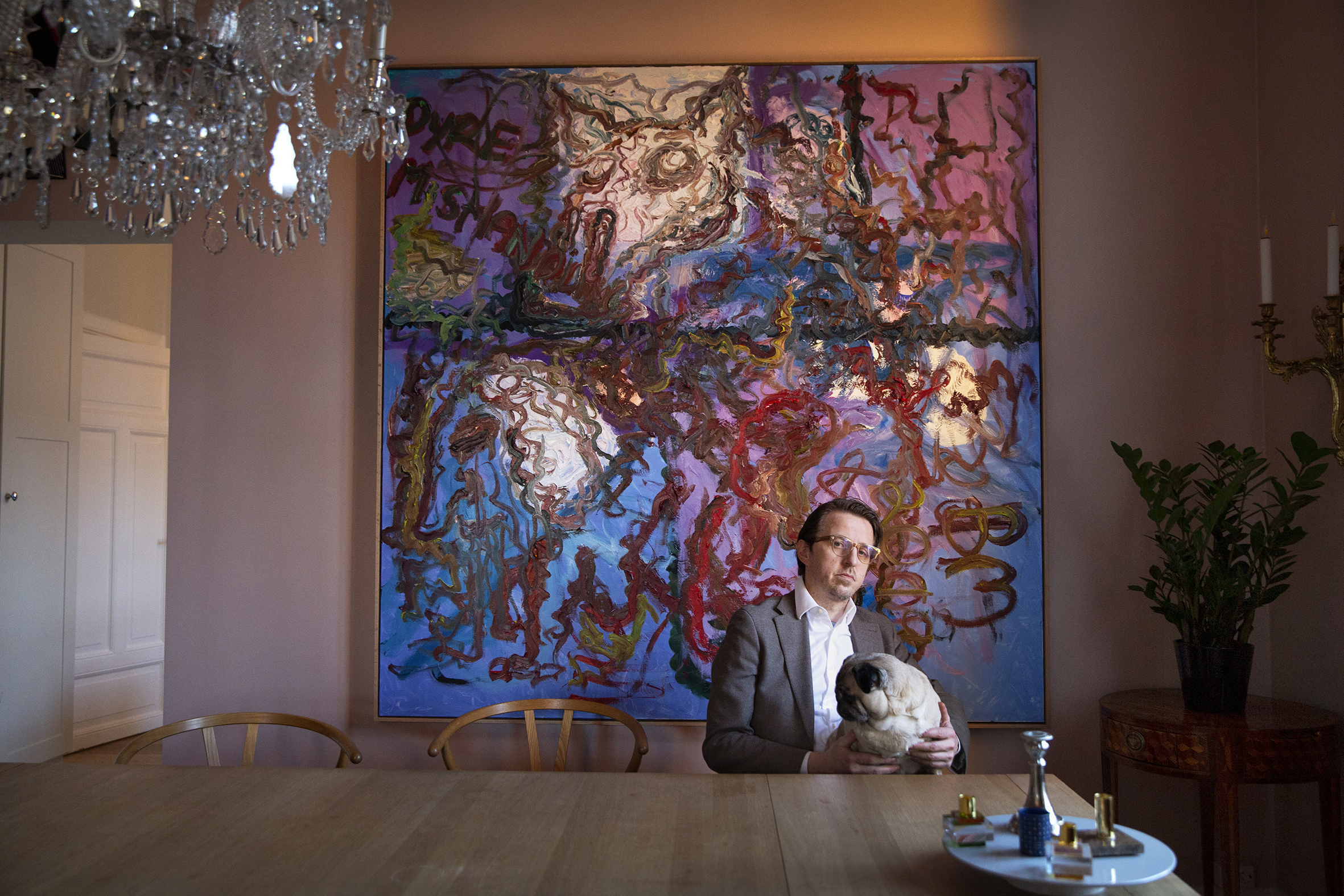 Sverre Bjertnæs sitter ved spisebordet i leligheten sin, bak han henger et stort maleri.