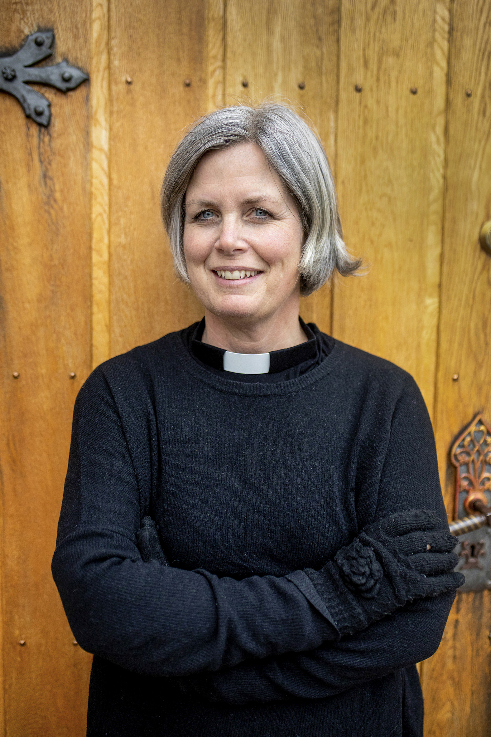 Sykehusprest Sunniva Johnsen Fisknes står med armene i kors utenfor kirkedøren. Prestekragen stikker opp fra halsen på ullgenseren hun har på seg. PÅ hendene har hun hanser. Hun smiler til kameraet.