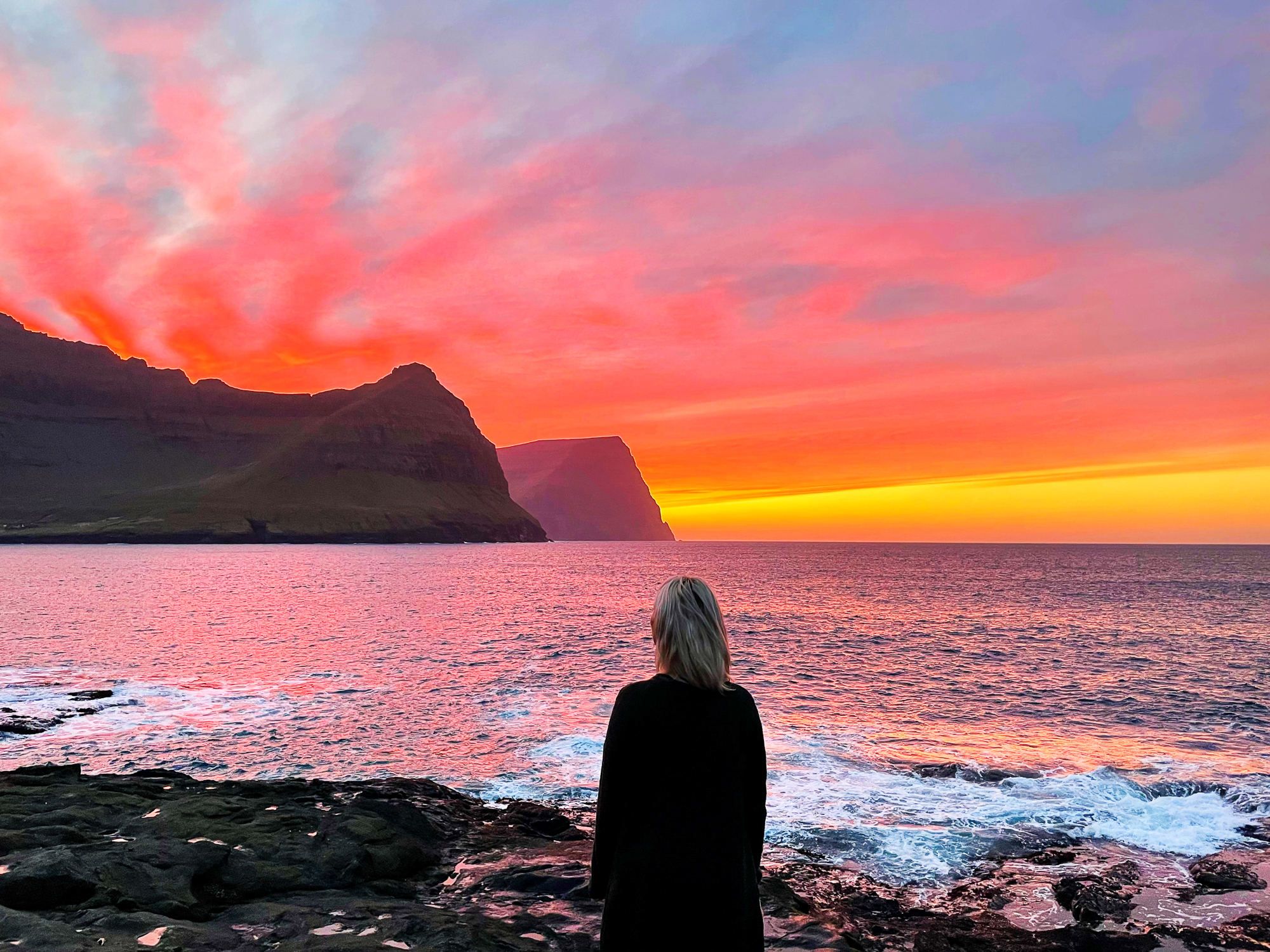 Kvinne med ryggen til som ser på en vakker solnedgang i havet, bak noen klipper.