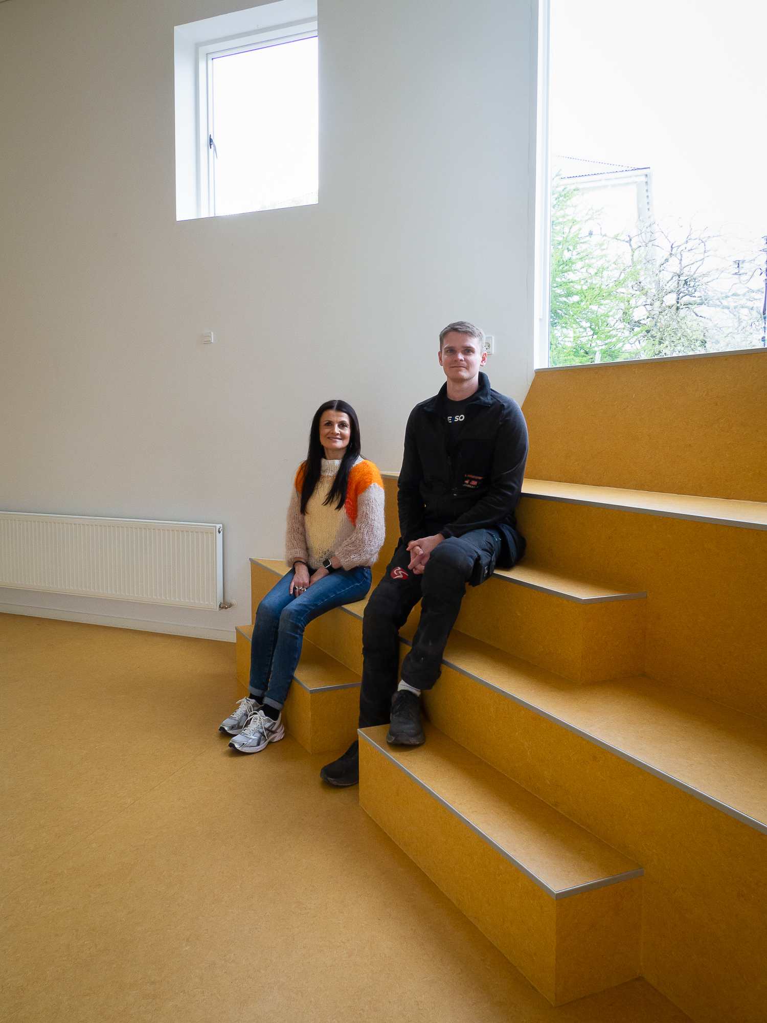 Ung gut og kvinne sitter på ulike trinn på en liten innendørs trapp man kan sitte på.