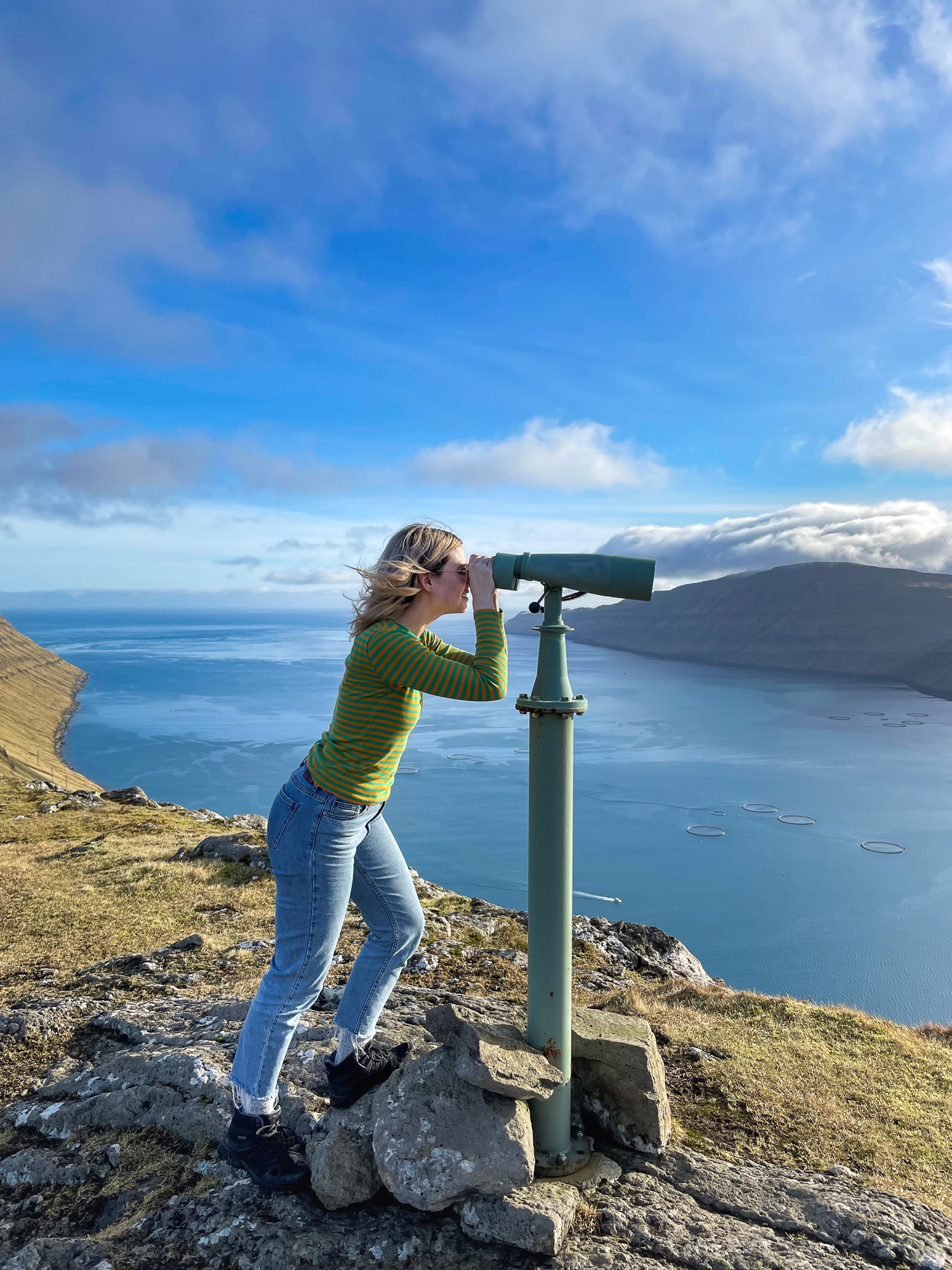 Ung kvinne som kikker utover hav og landskap i en fastmontert, stående kikkert oppå fjellet.