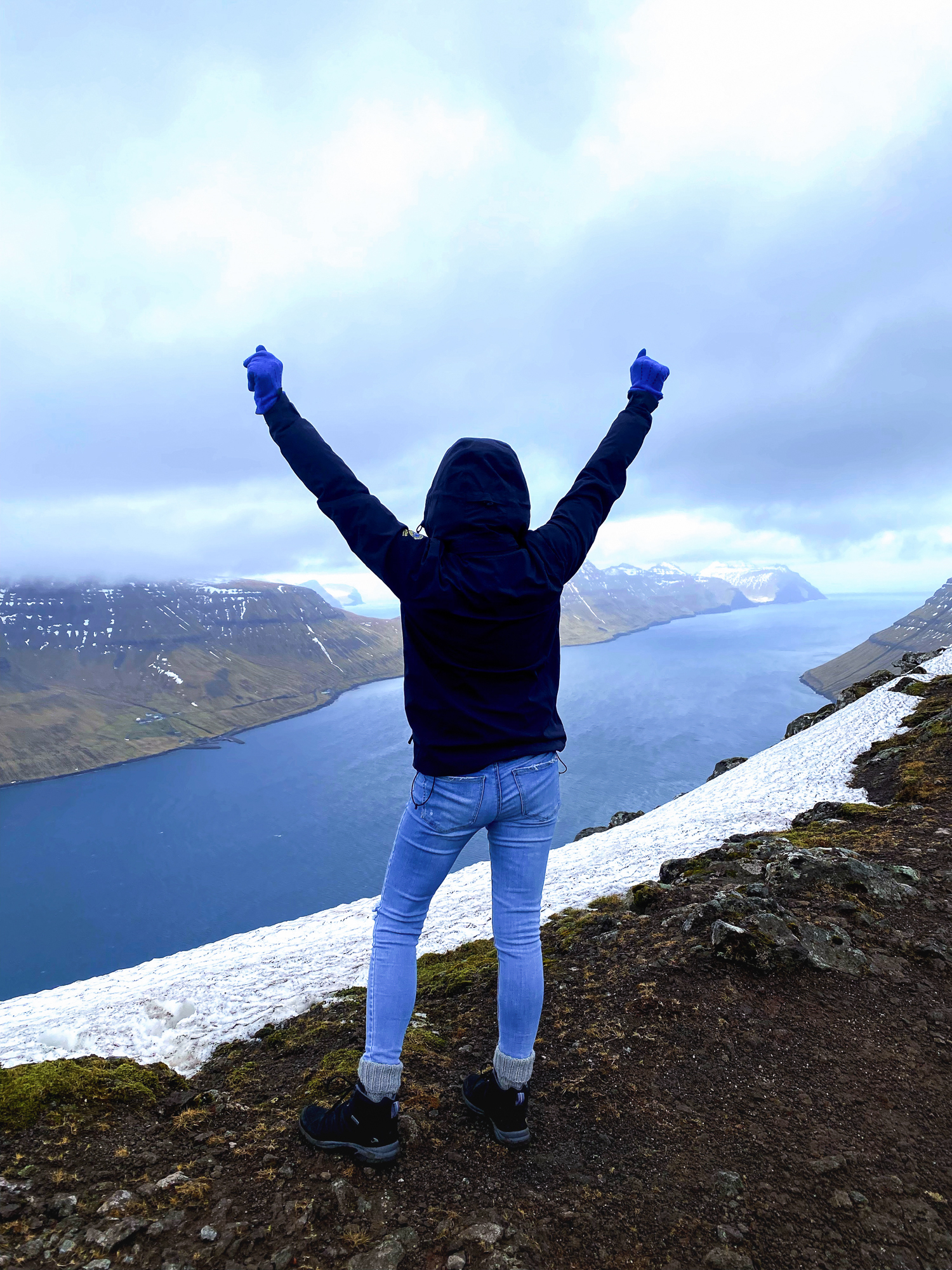Kvinne som står på kanten av et fjell med ryggen til og begge armene opp i været. Hun ser utover en fjord.