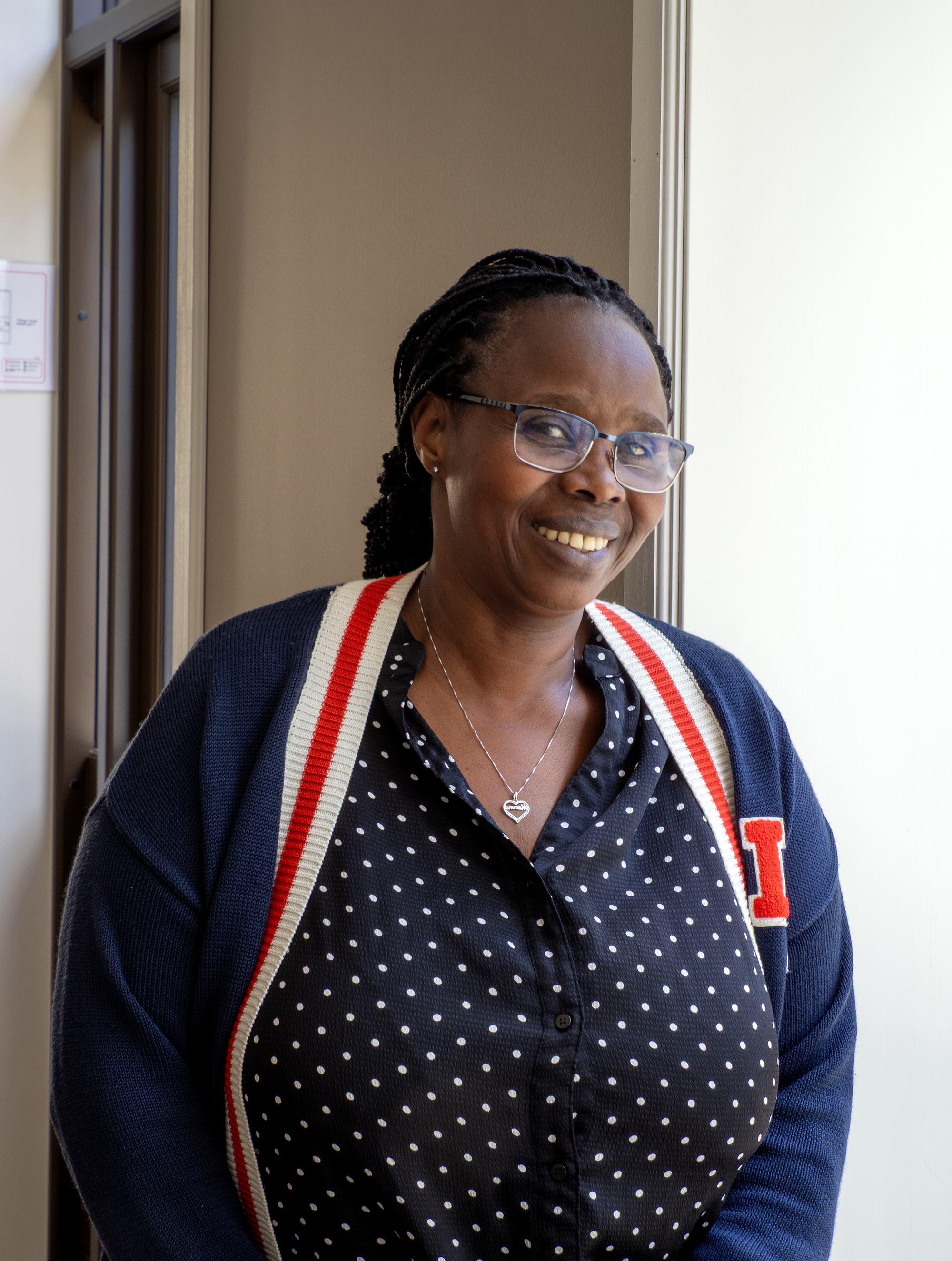 Foto: Svart kvinne med briller og fletter står ved et vindu. Hun har på seg blå bluse med hvite prikker og en strikkejakke i blått med striper langs åpningen i hvitt og rødt.