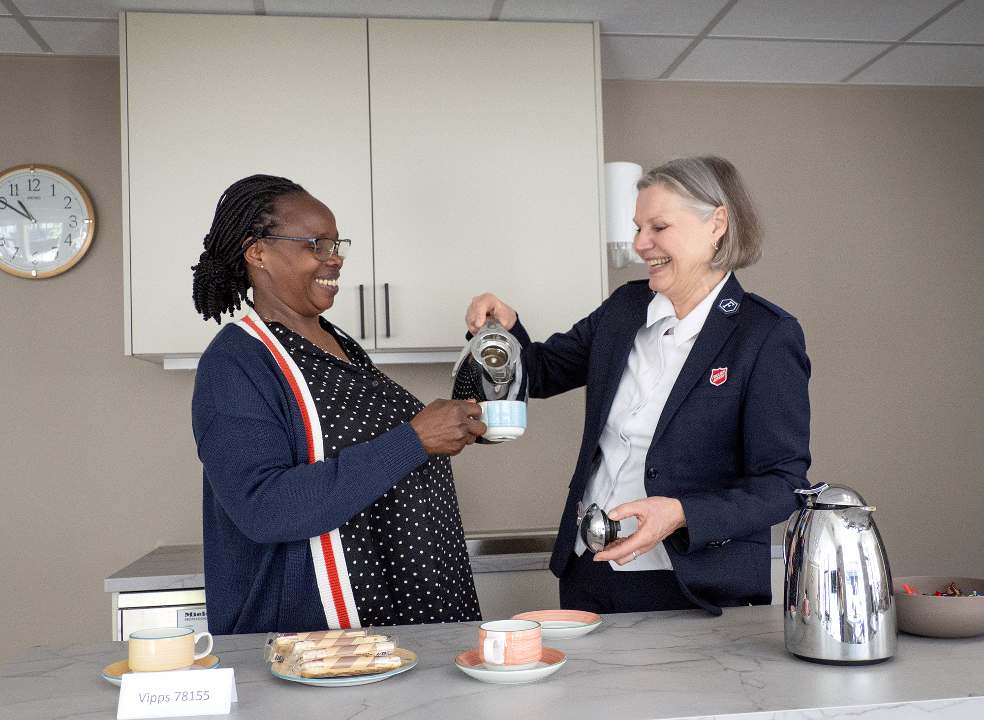 Foto: En svart og en hvit kvinne skravler over skjenking i en kaffekopp på et hvitt kjøkken.