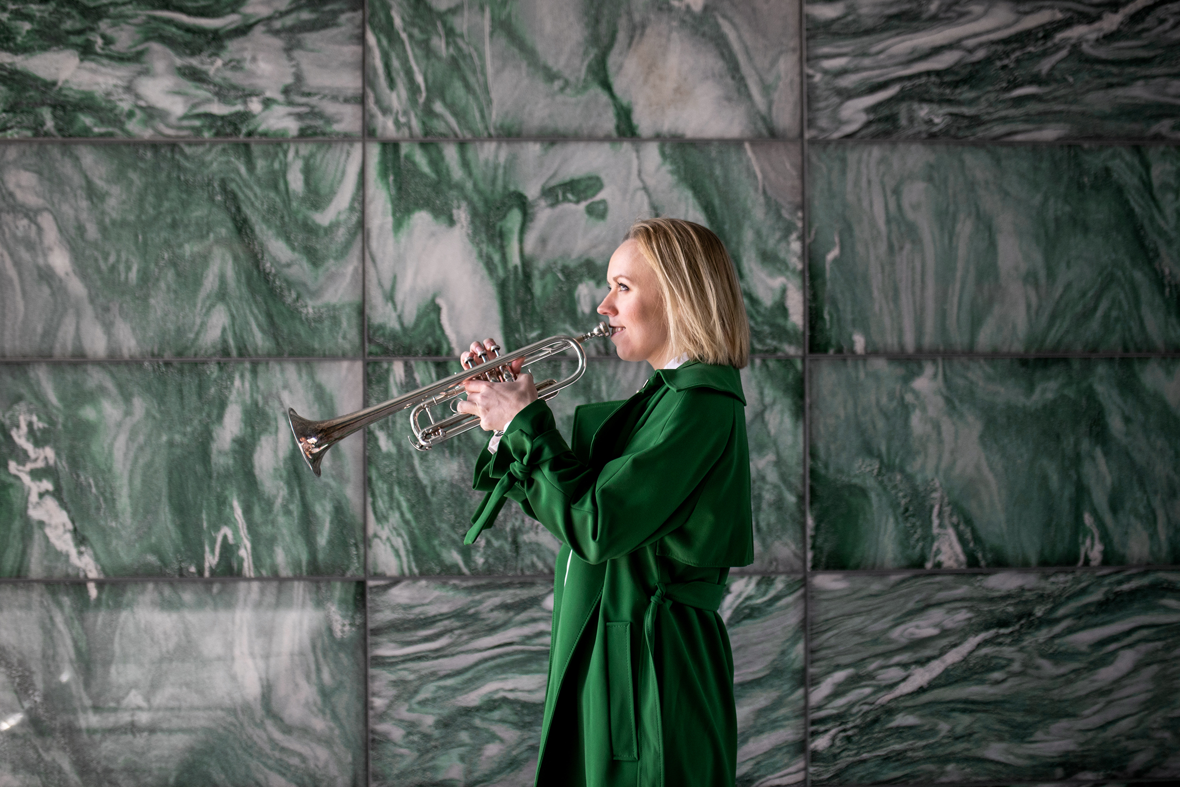 Tine, i grønn kåpe, står i profil foran en grønn marmorvegg og spiller på trompeten