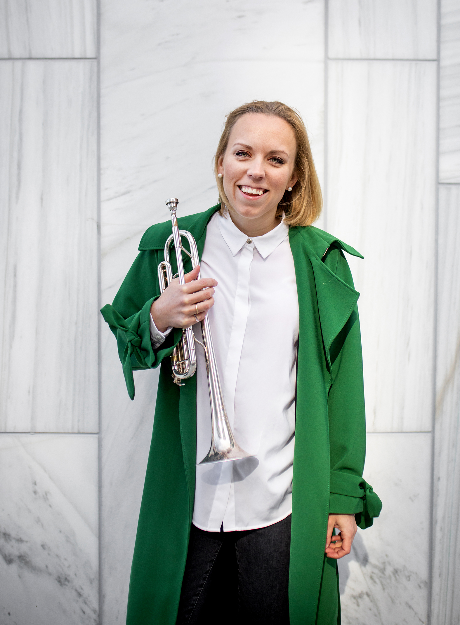 Tine står foran en hvit marmorvegg i grønn kåpe med trompeten i hånden