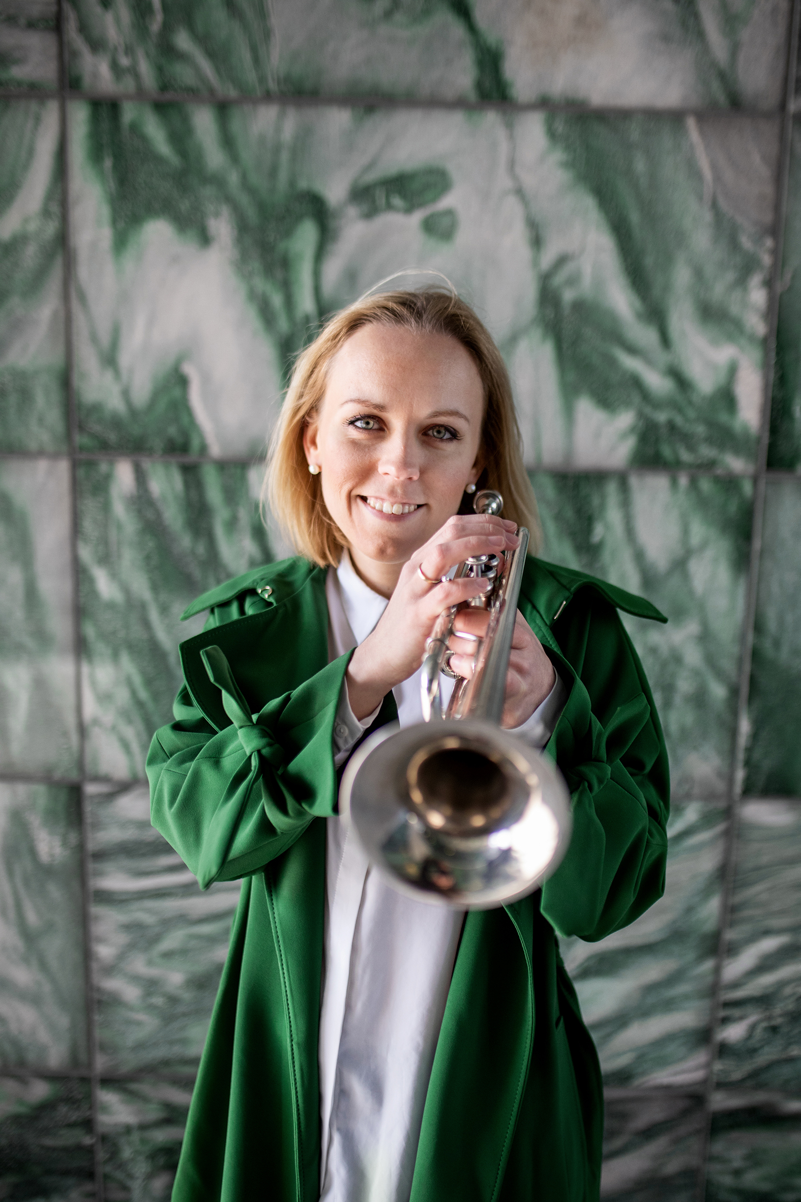 Tine står foran en grønn marmorvegg og ser rett i kamera mens hun spiller på trompet
