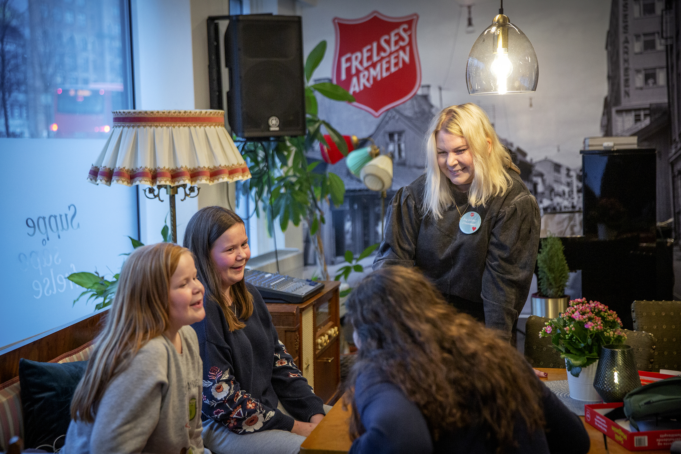Tone Berg står og smiler og snakker med flere personer som sitter ved et kafebord på Sammen.