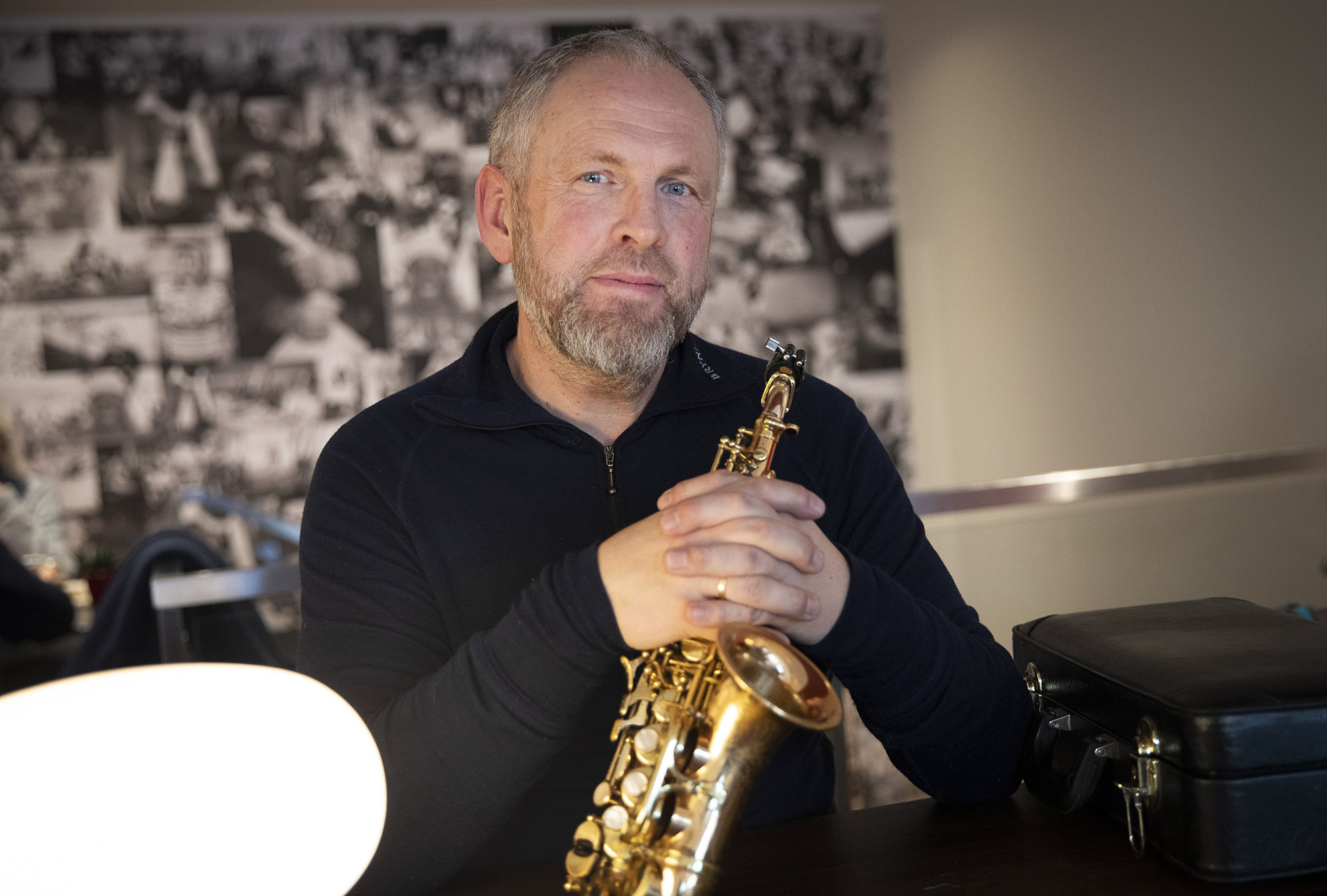 Foto: Mann med saksofon sitter på en kafé. Han har gråblondt hår og skjegg.