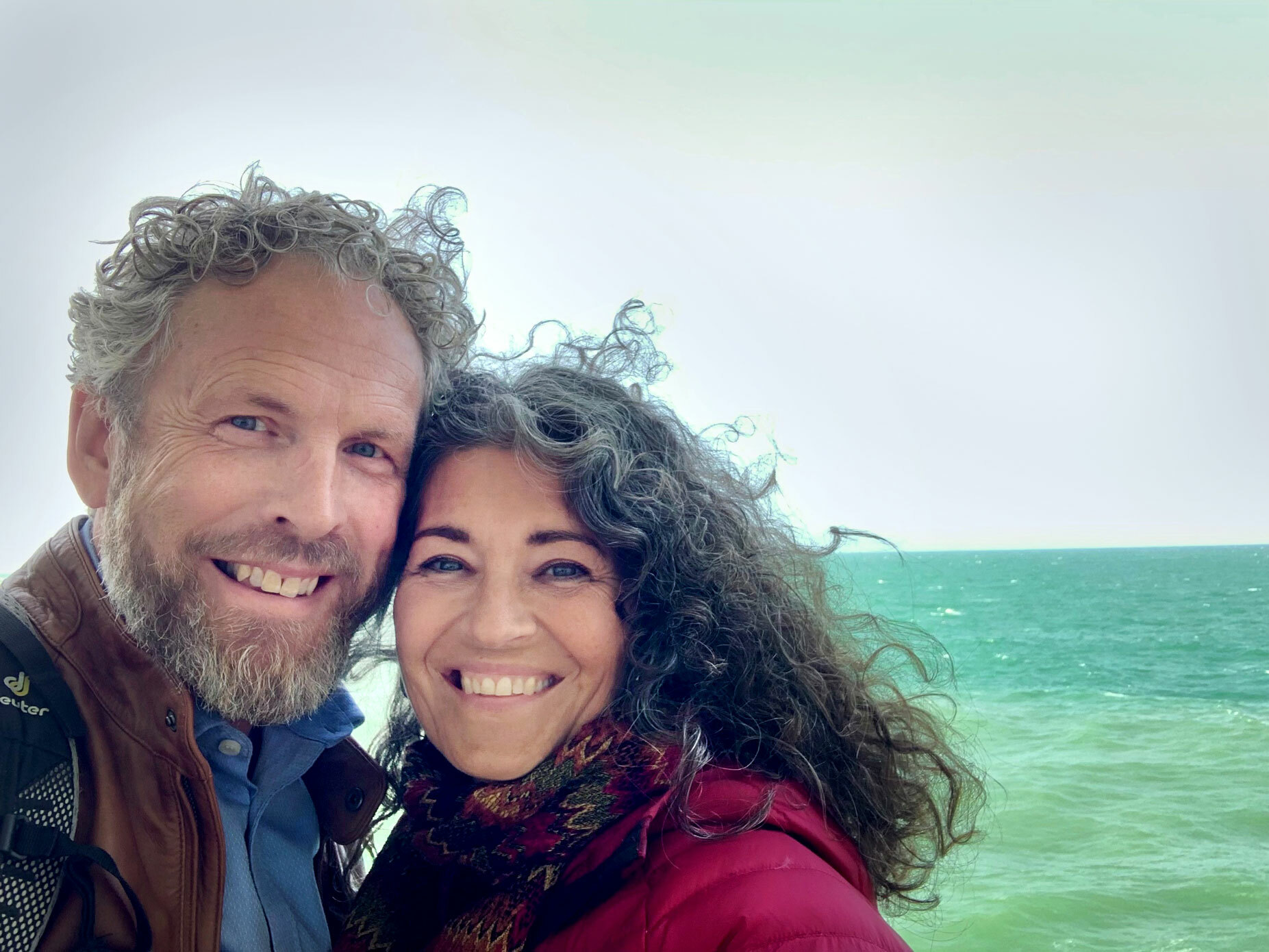 Foto: Mann og kvinne i 50-årene står kinn mot kinn. Bak dem er det grønt hav og vinden blåser i krøllene til begge. Hun er mørk, han gråblond med skjegg.