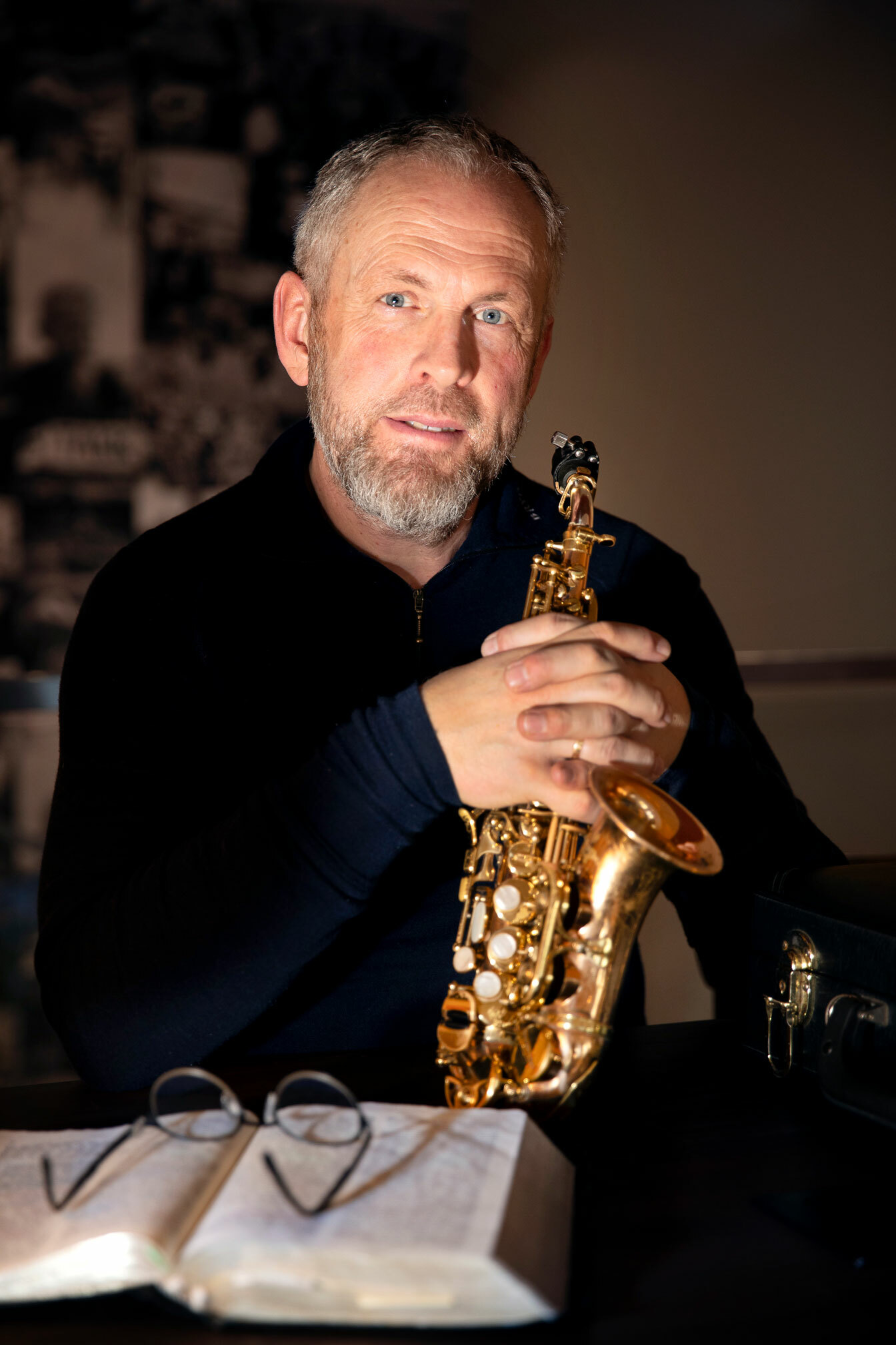 Foto: Mann sitter inne på en kafé med en saksofon- Han folder hendene over den og foran seg har han en tykk bok og oppå den ligger et par lesebriller.
