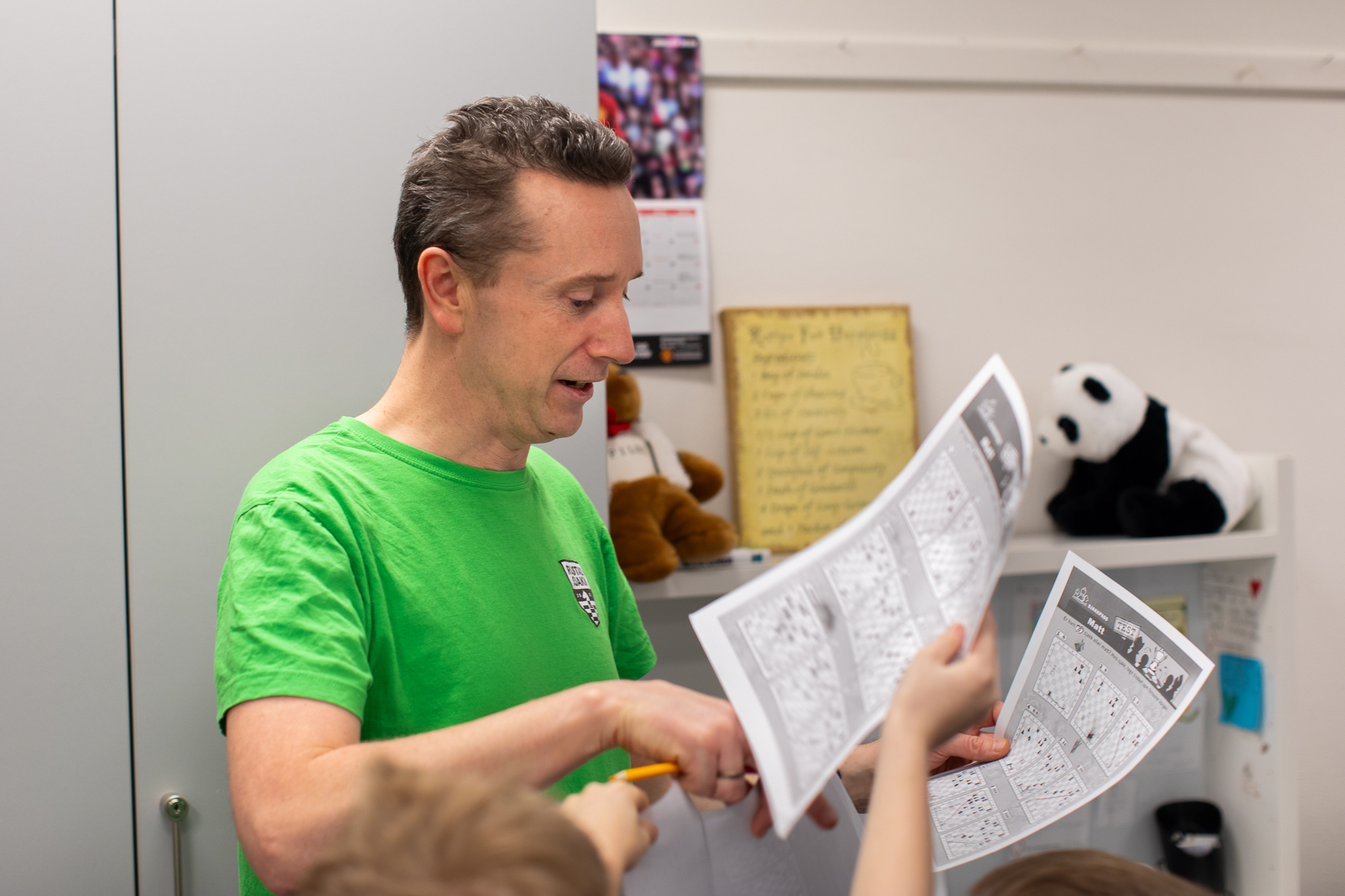 Flere barnehender rekker et ark med sine svar i retning en mann i grønn T-skjorte som står inne i et klasserom.